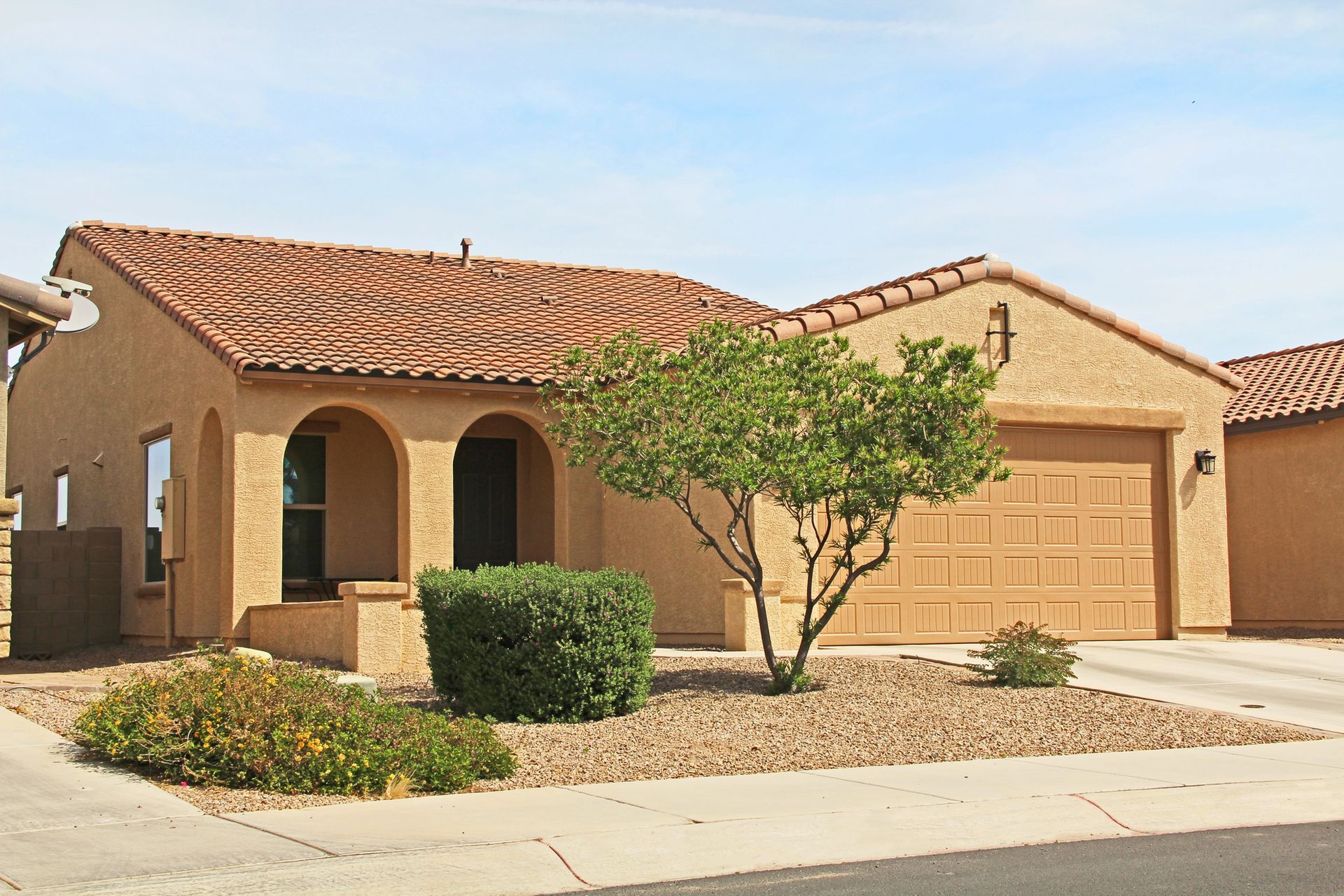 Tan stucco house with terracotta roof, arched entry, and tan garage door; gravel landscaping.