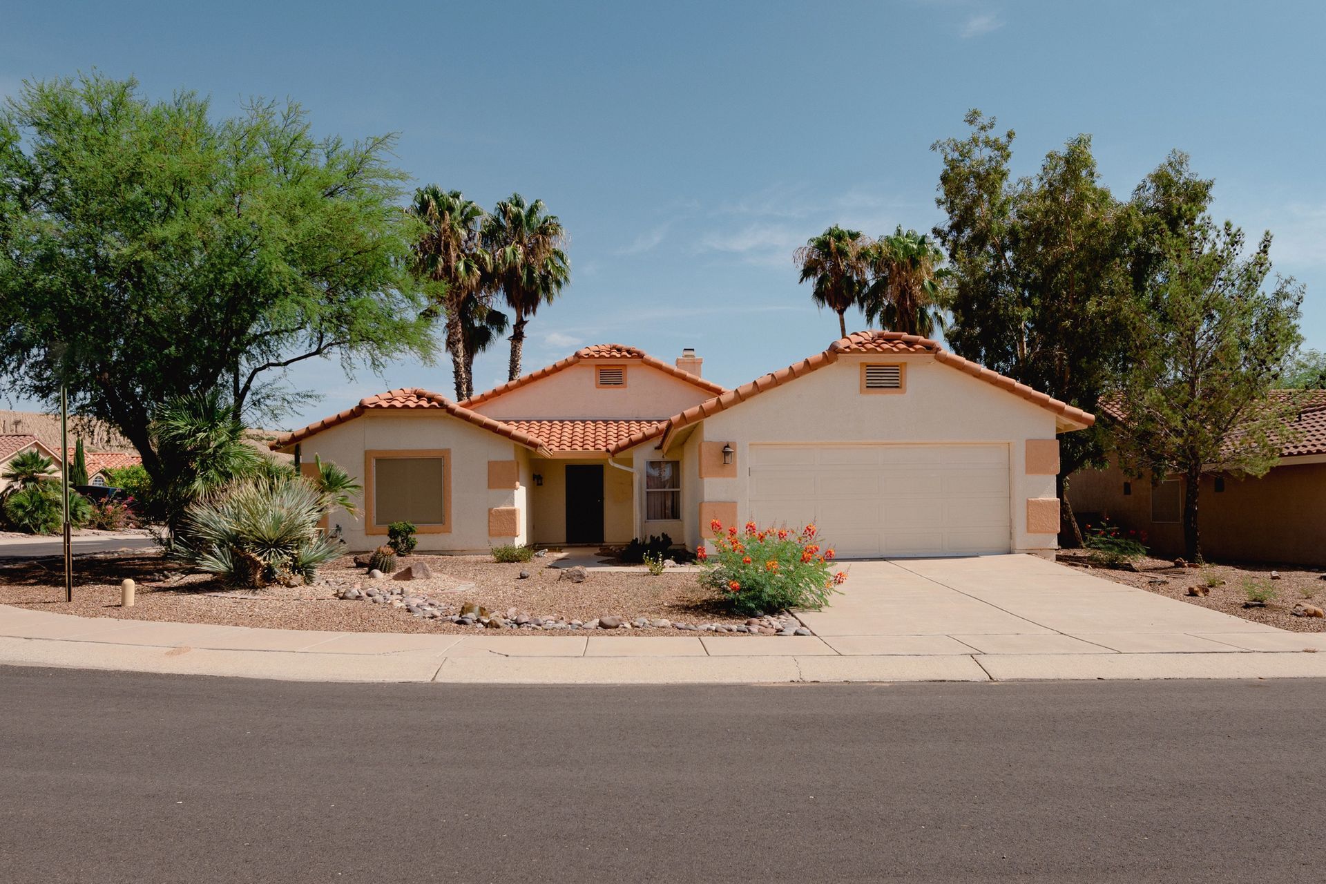 Tan house with red-tiled roof and two-car garage under a blue sky, surrounded by palm trees and desert landscaping.
