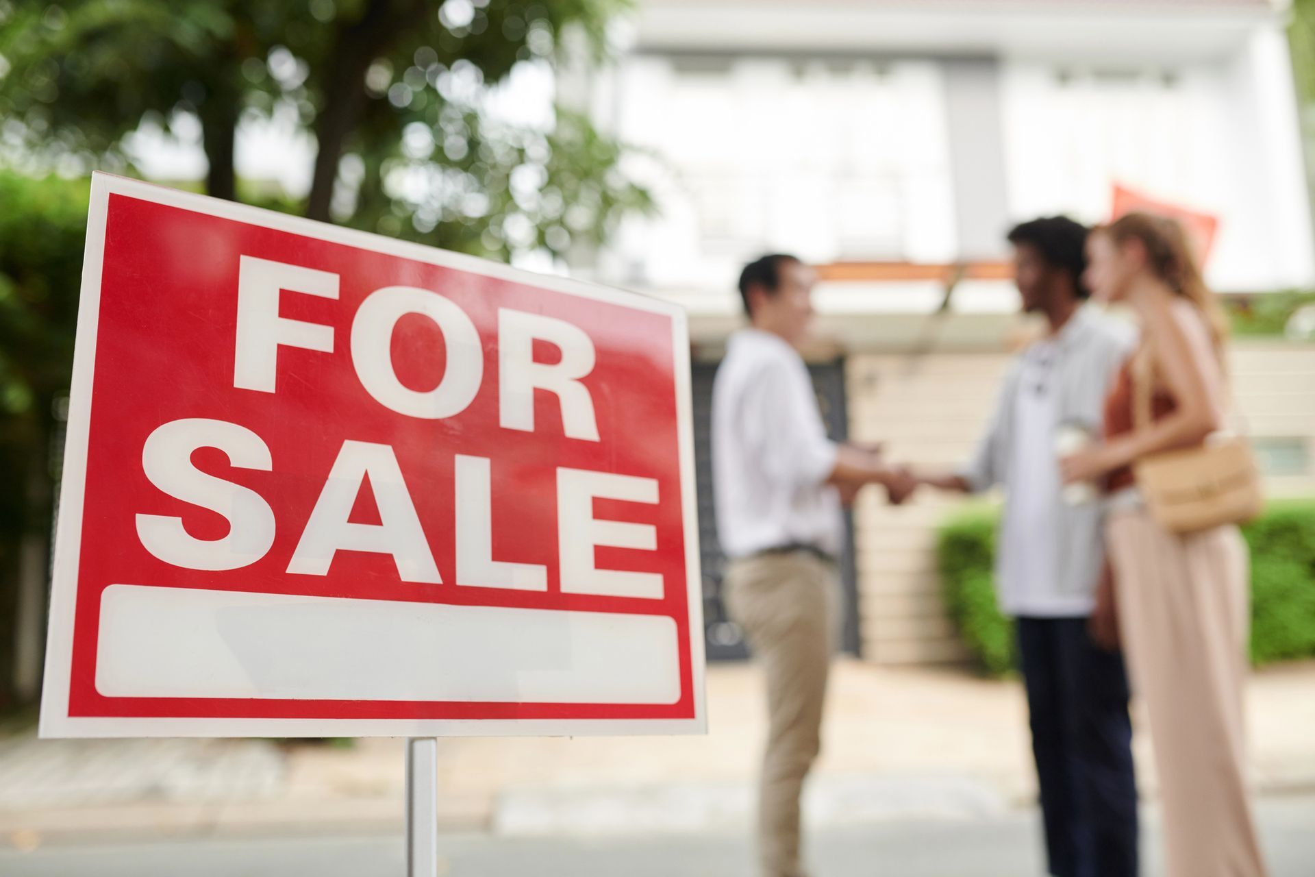 Red FOR SALE sign in front of a house, with a realtor shaking hands with a couple.
