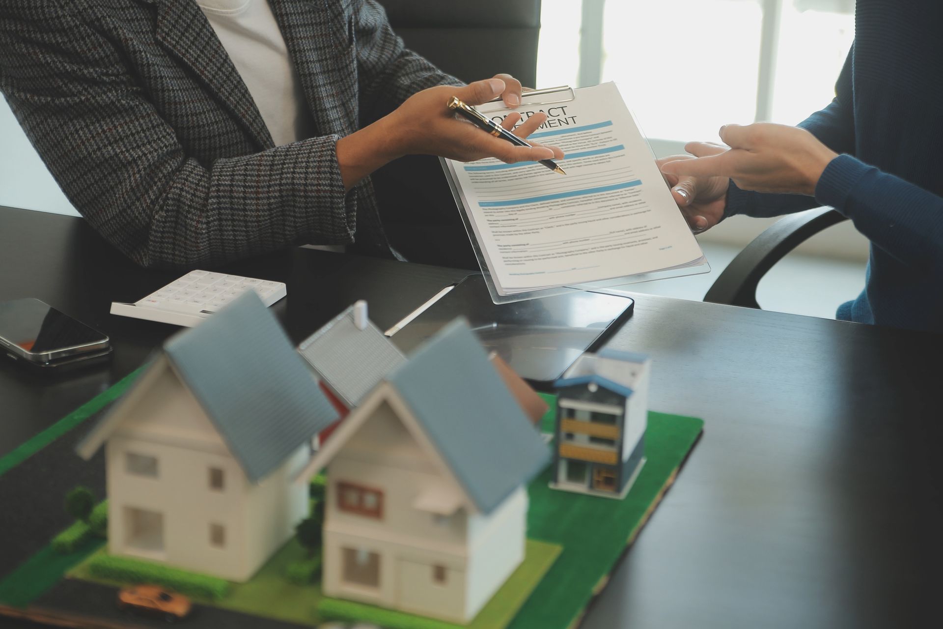 Person handing a document to another person; model houses on a desk.