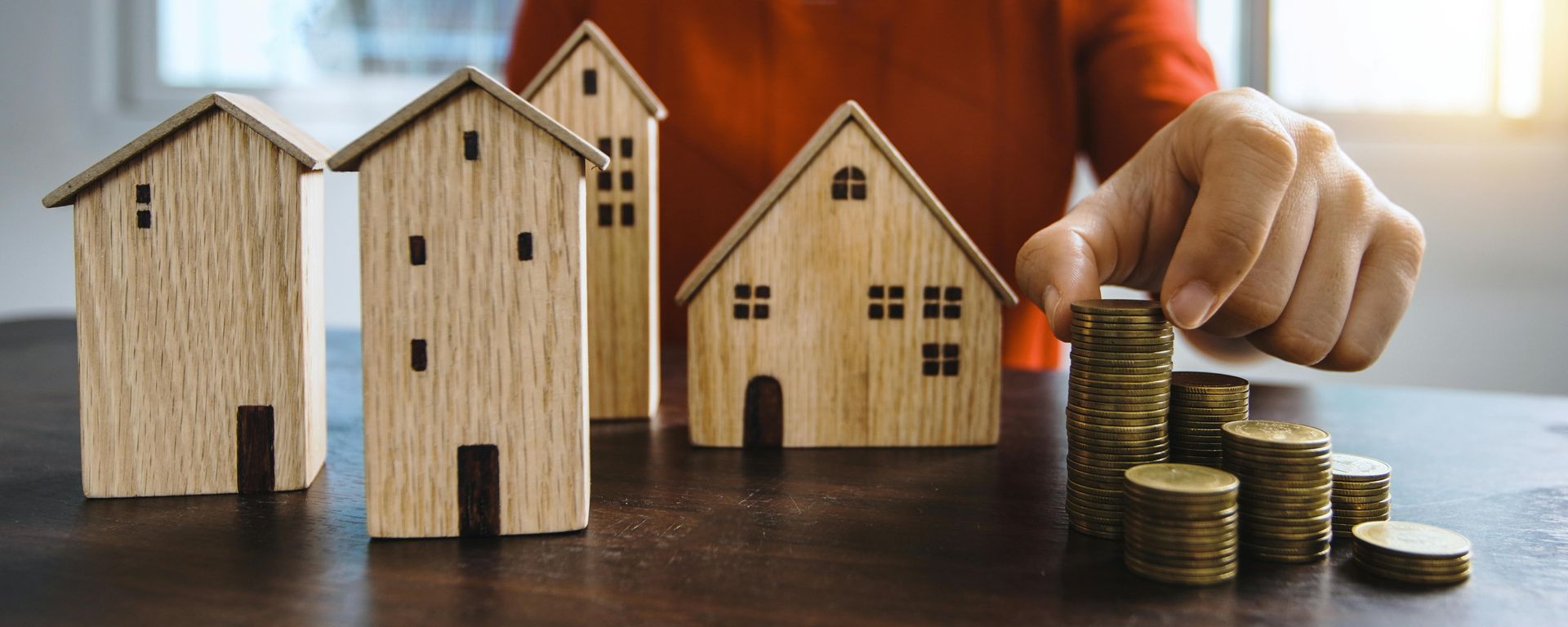 Hand stacking coins near wooden houses, symbolizing real estate investment or savings.
