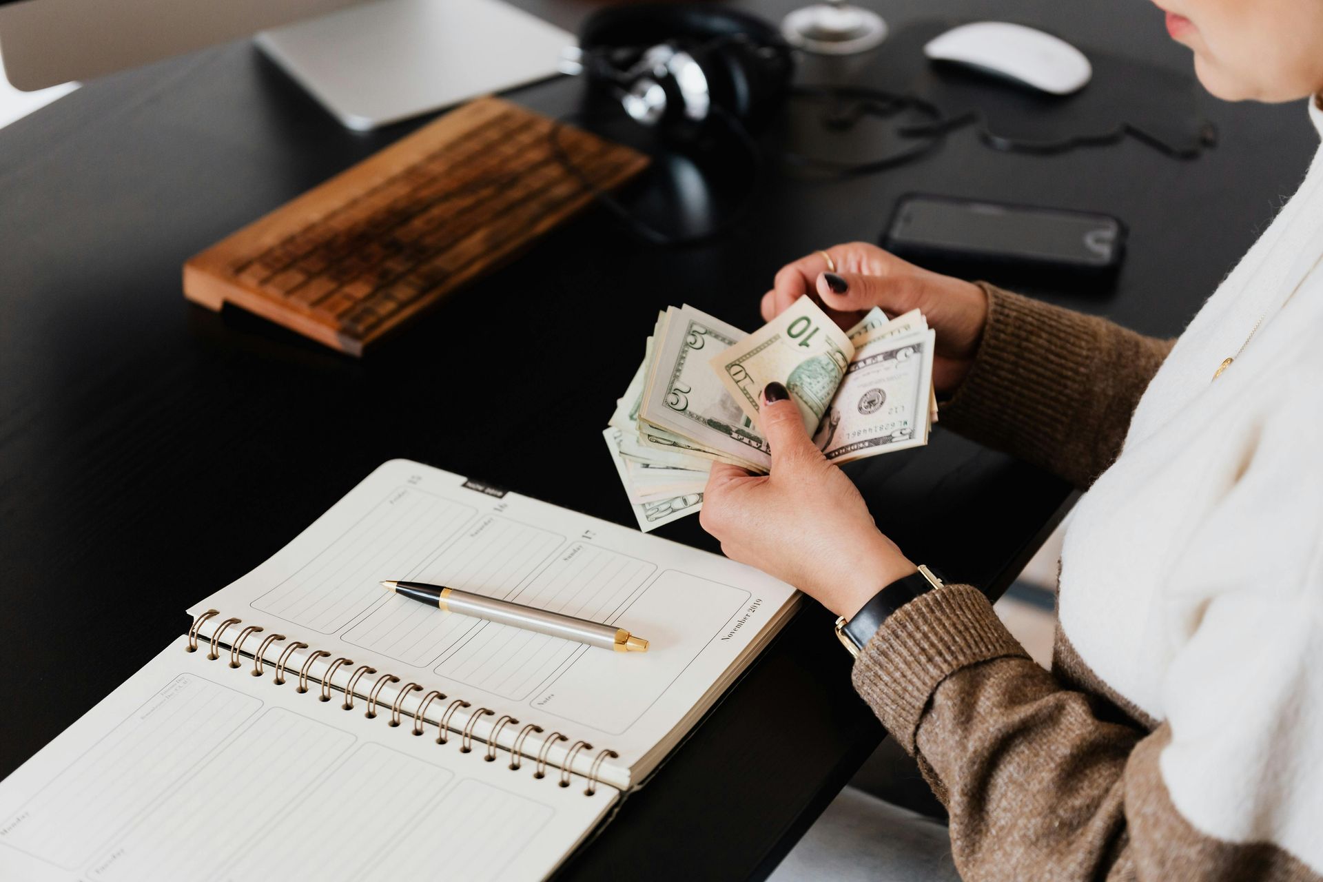 Person counting money at a desk with a notebook, pen, phone, and headphones.
