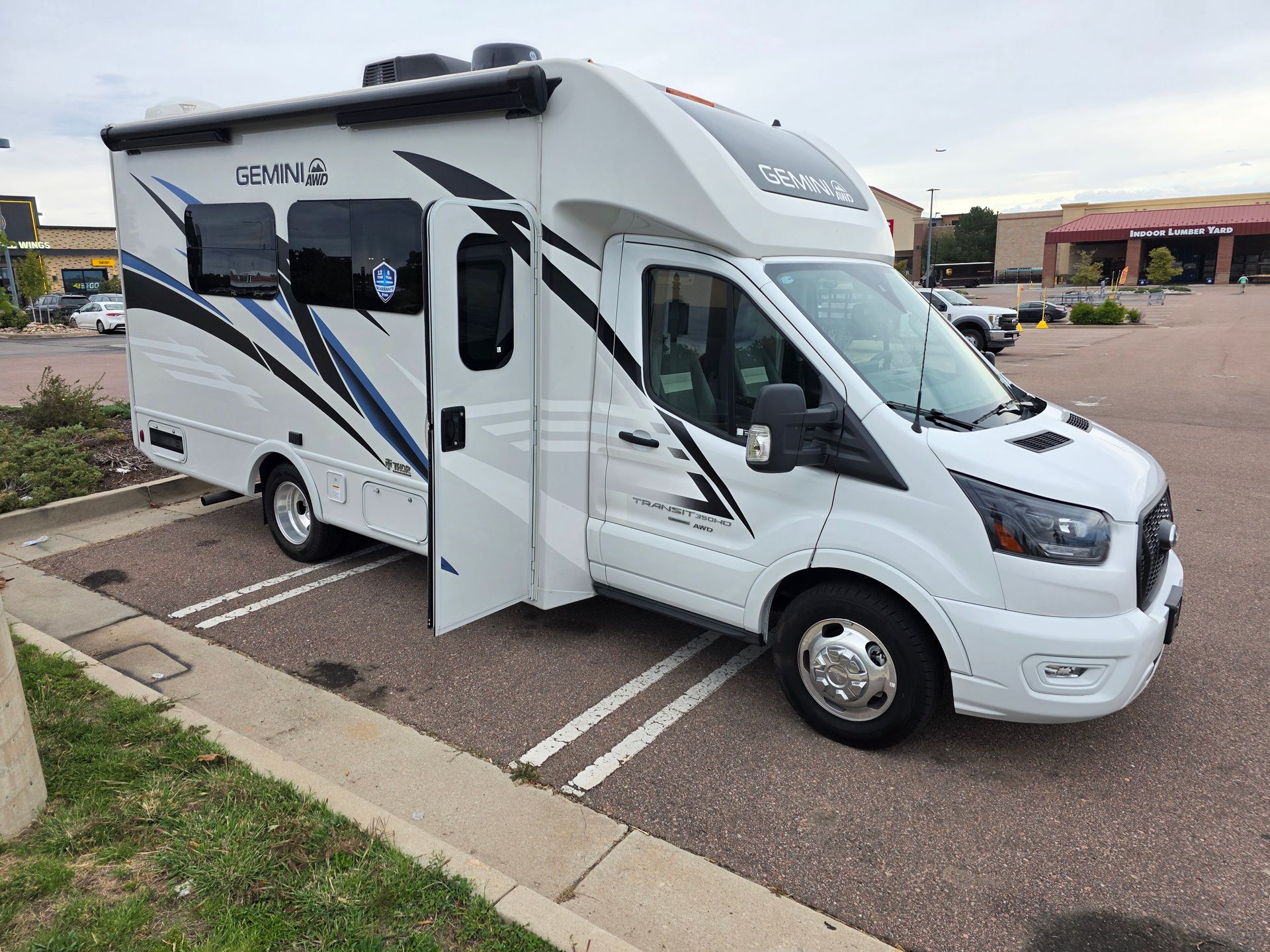 White Ford Thor Gemini RV parked outdoors under a blue sky. The RV has an awning and graphics.