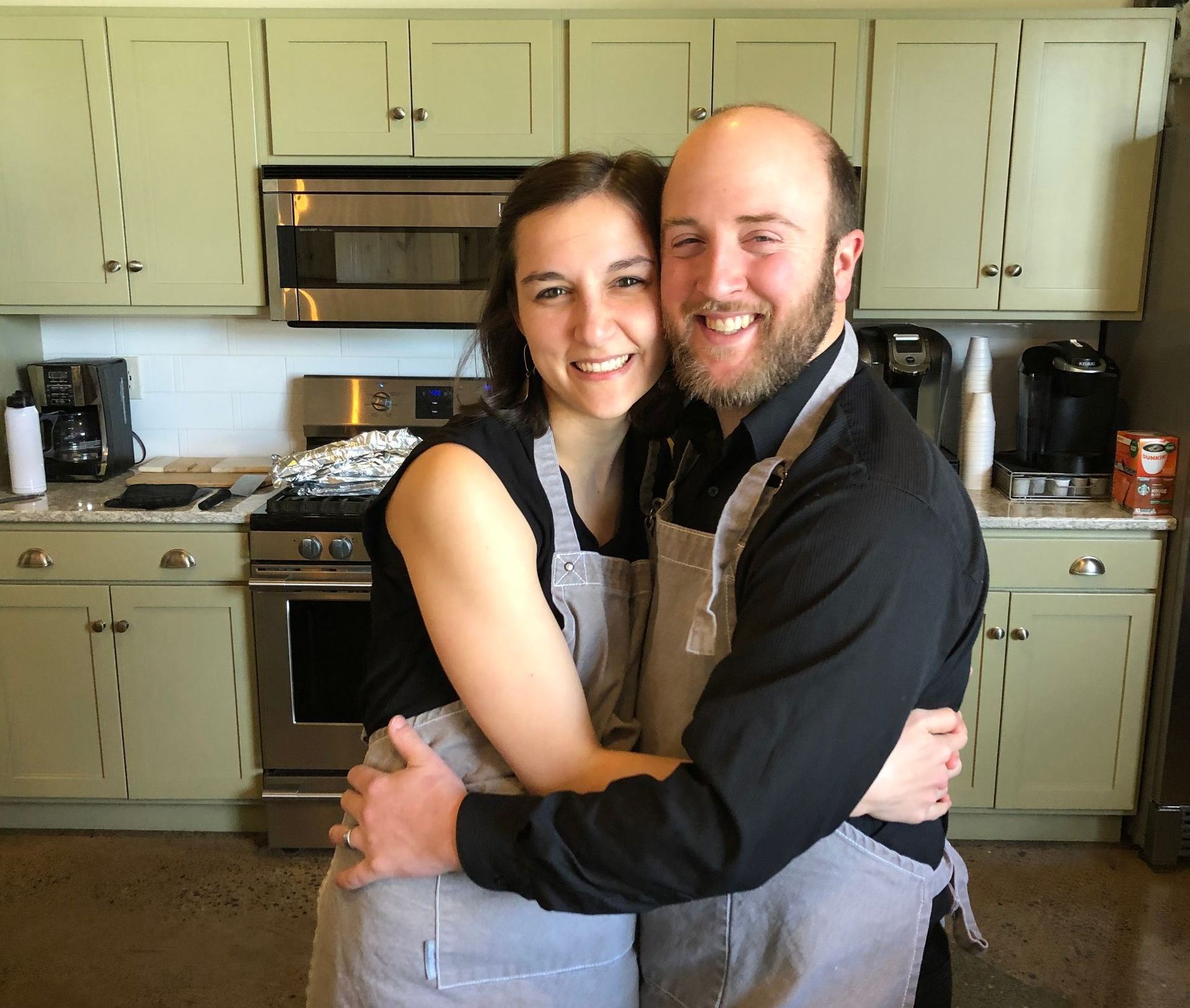 Seth and Mary Martin hugging in a kitchen wearing aprons