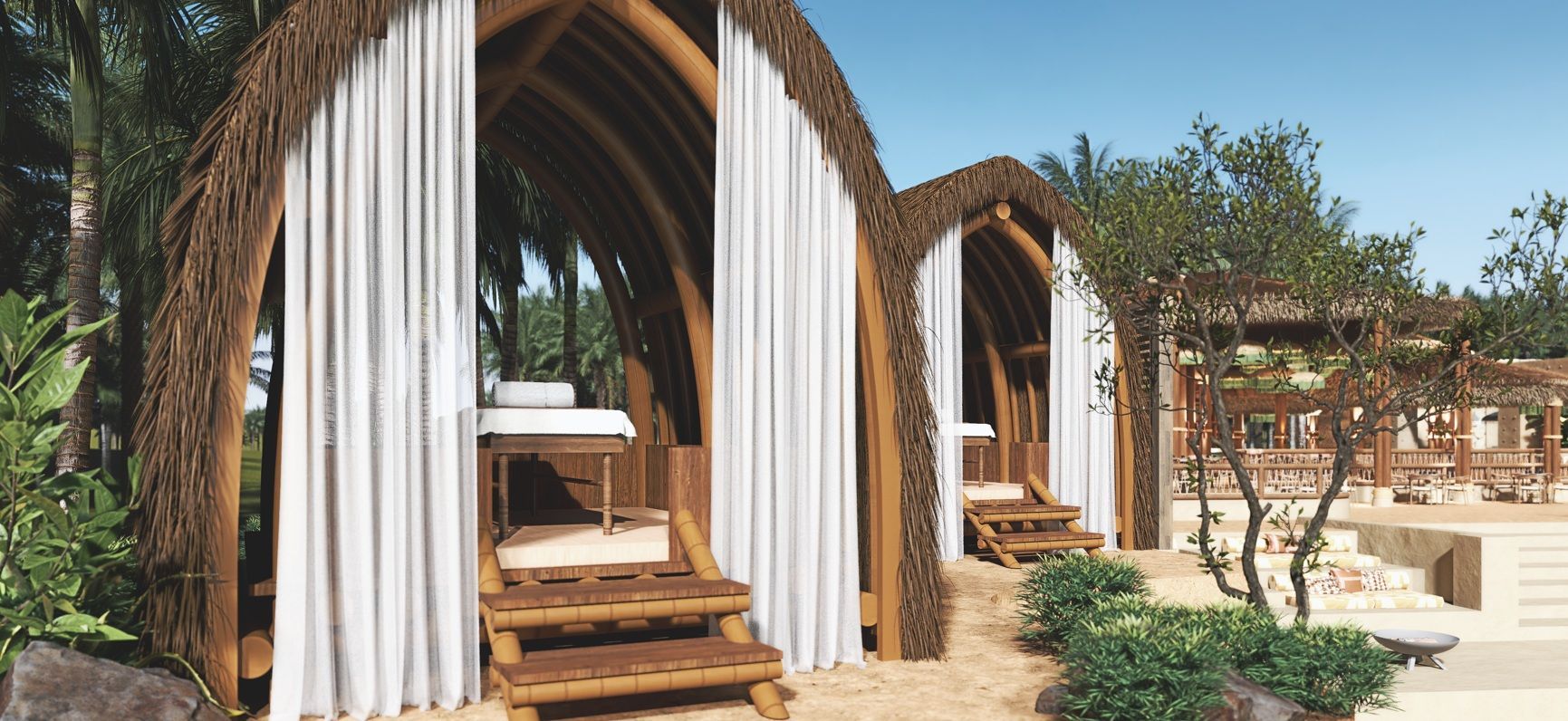 Tropical spa huts with white curtains, a massage table, and wooden steps on a sandy beach under a clear blue sky.