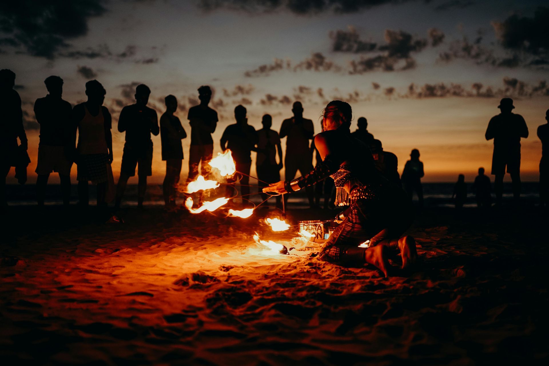A group of people are standing around a fire on the beach at night.