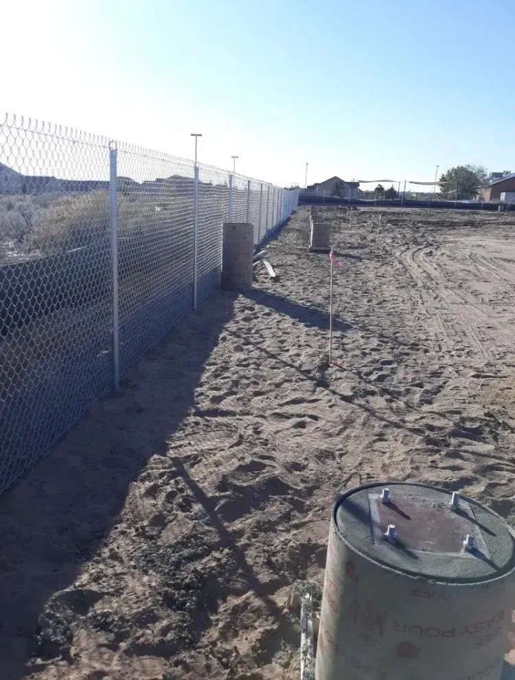 A chain link fence surrounds a dirt field with a barrel in the foreground.