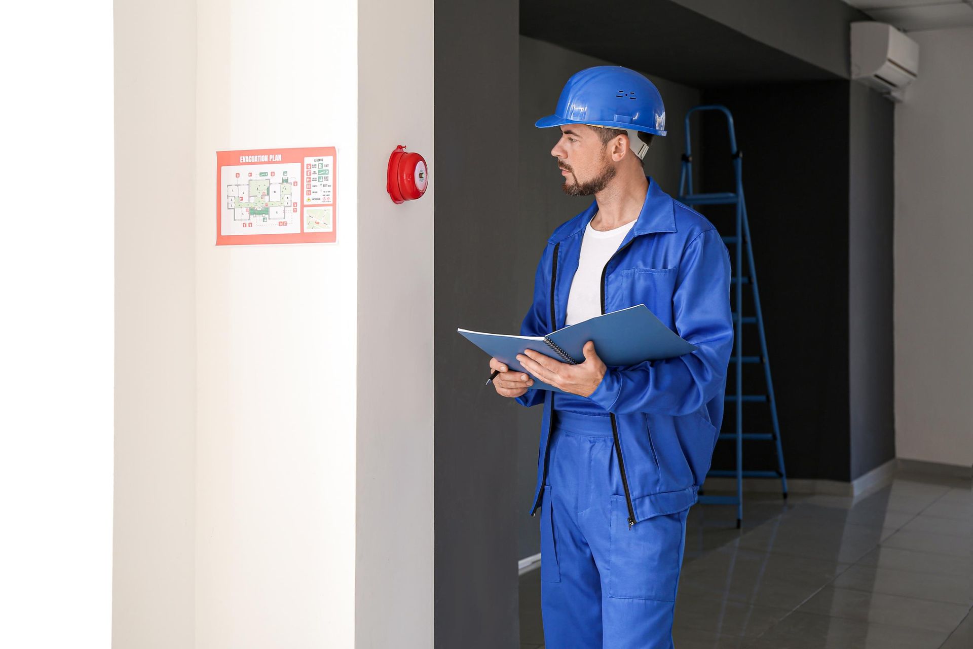 A man in a blue uniform is holding a clipboard and looking at a fire alarm.