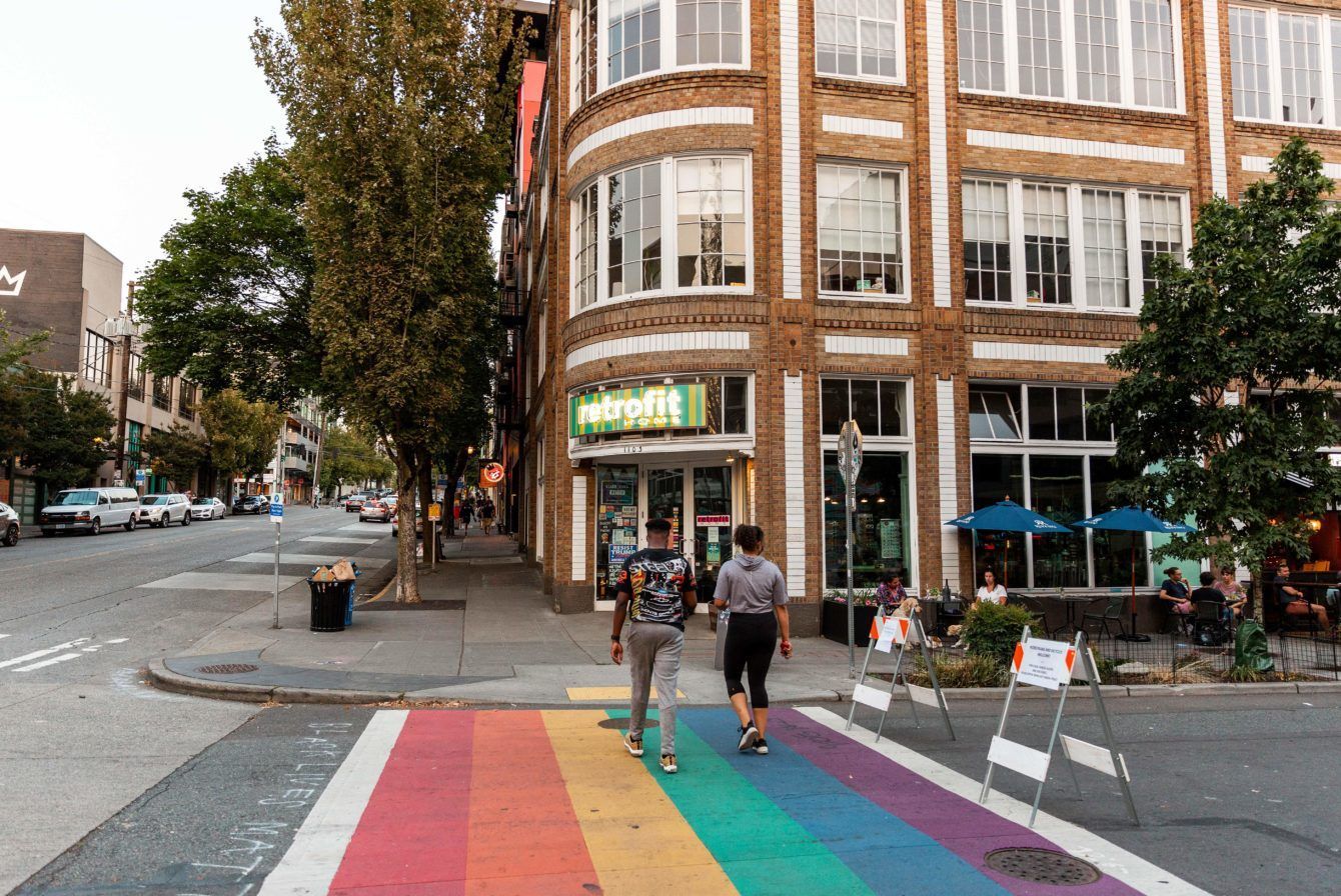 Two people are walking across a rainbow crosswalk in front of a building.