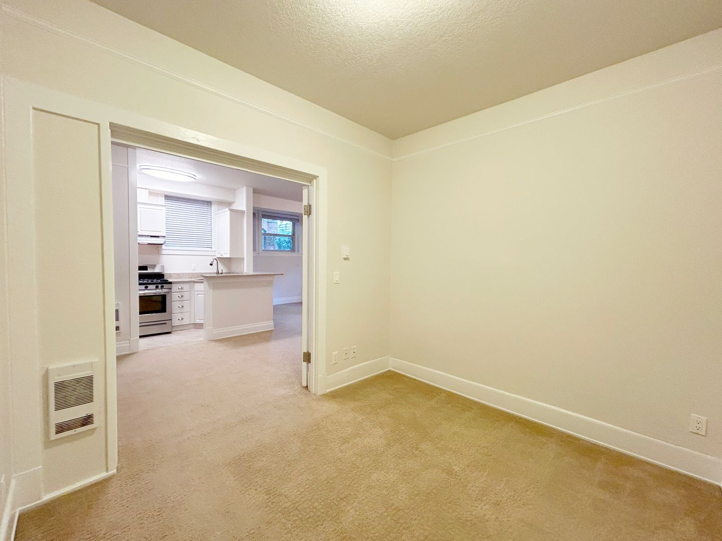 An empty room with a carpeted floor and a kitchen in the background.