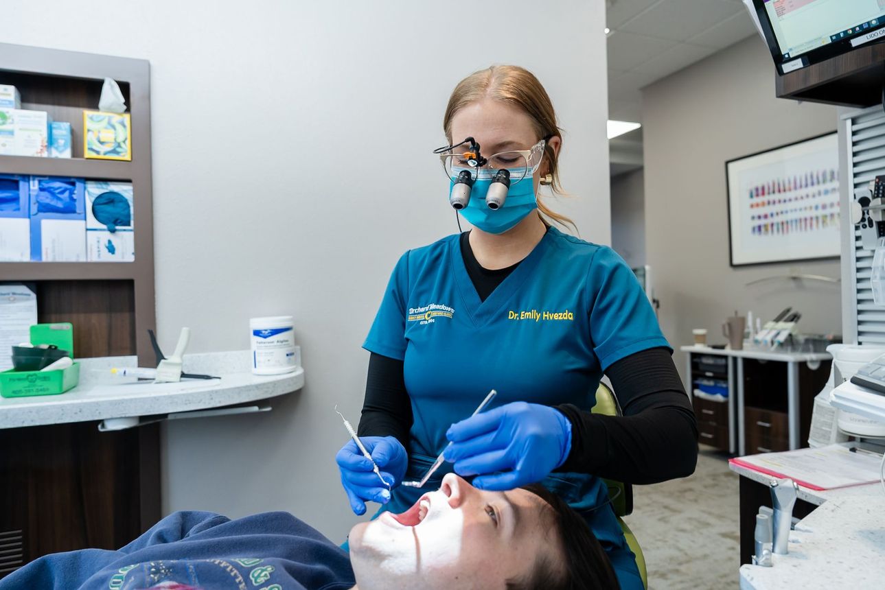 Dr. Emily in teal scrubs and loupes examines a patient in a clinical office setting.