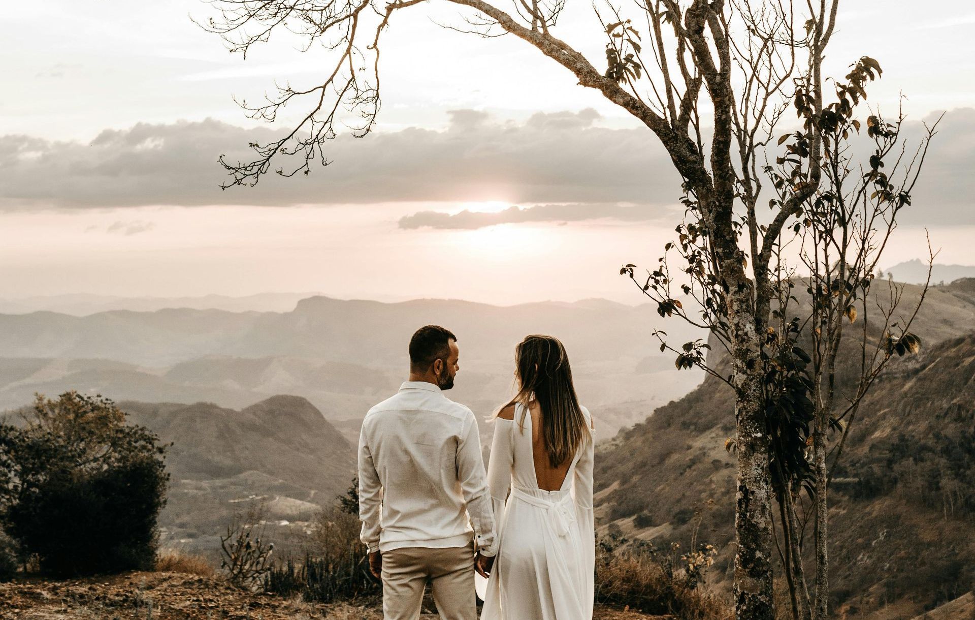 A man and a woman are standing next to a tree looking at the sunset.