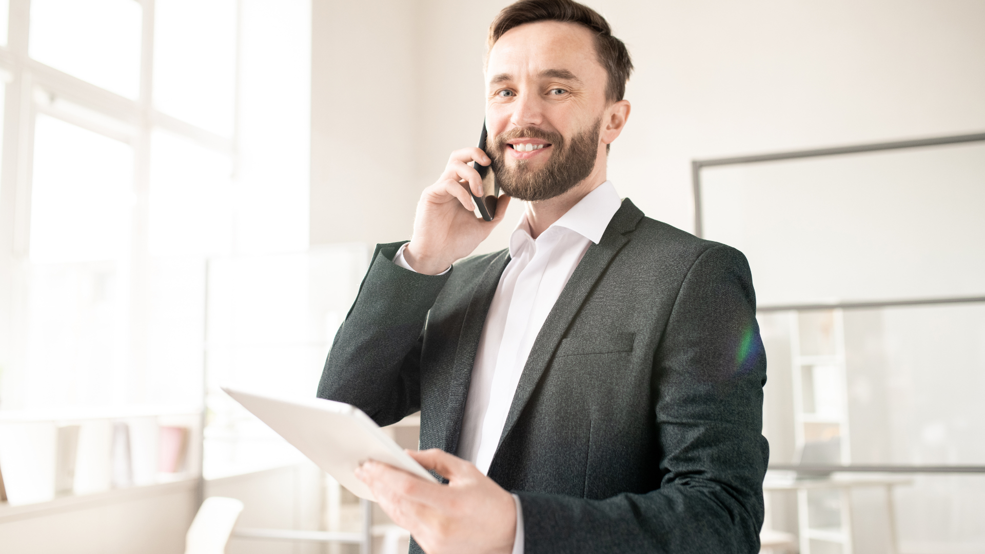 Man in suit, smiling while talking on a phone and holding a tablet in a bright office.
