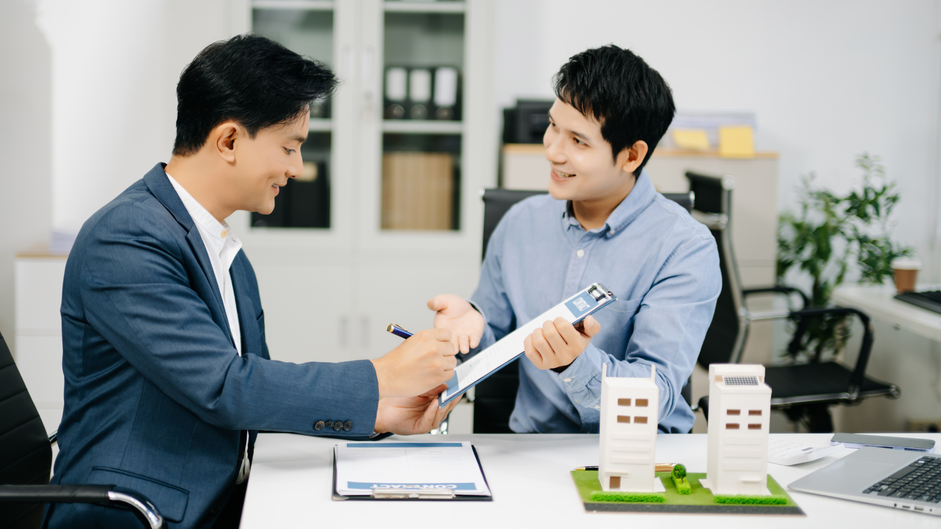 Two businesspeople smiling and shaking hands across a desk with model buildings and documents.