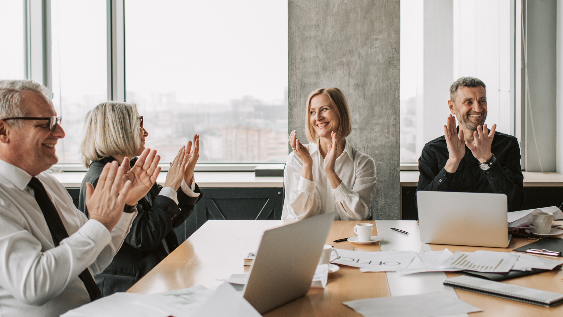Business team clapping in a conference room. Smiling people at a table with laptops, documents, and coffee cups.
