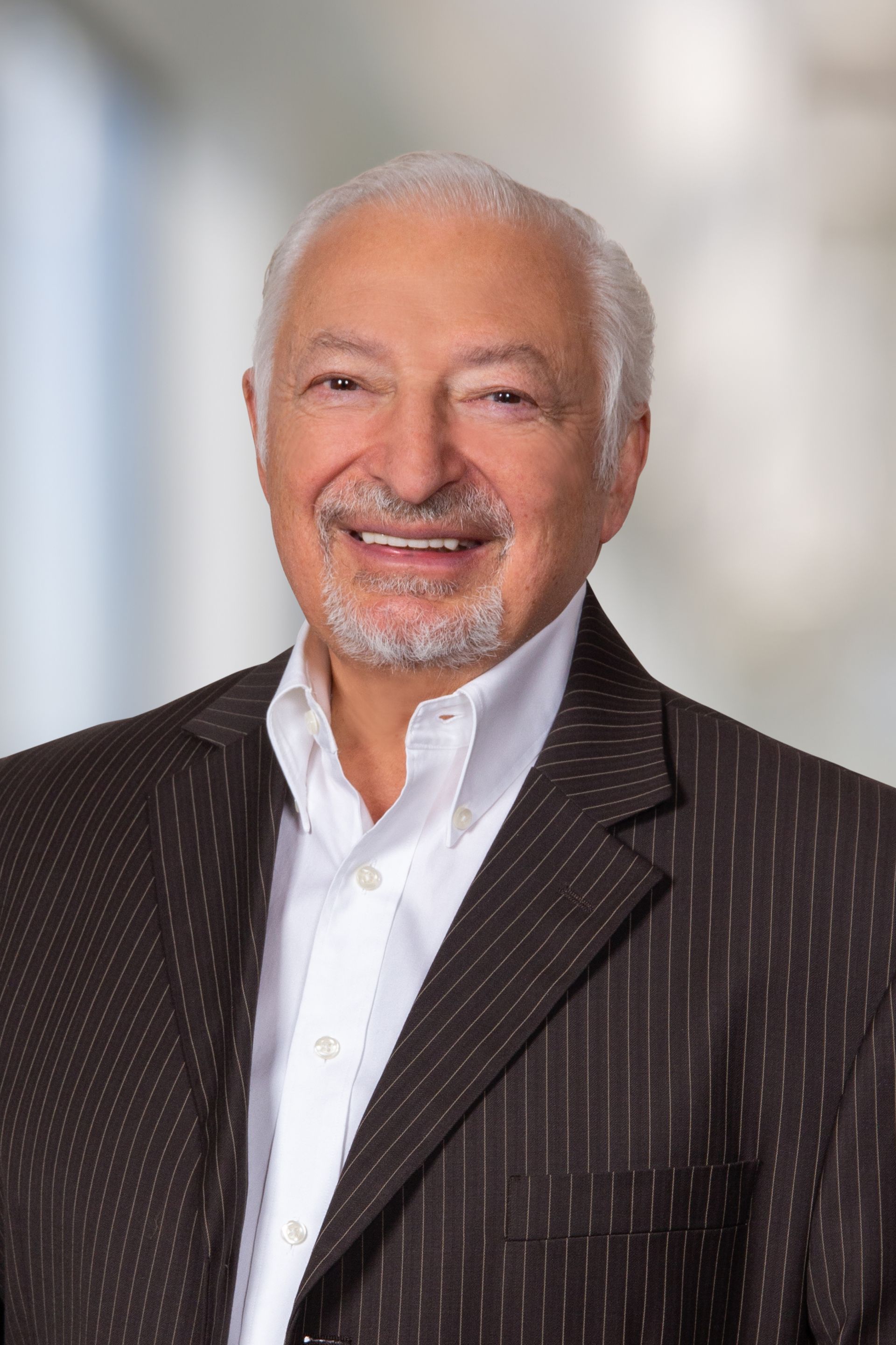 Smiling older man in a brown pinstripe suit jacket and open-collared white shirt, posing in front of a blurred background.