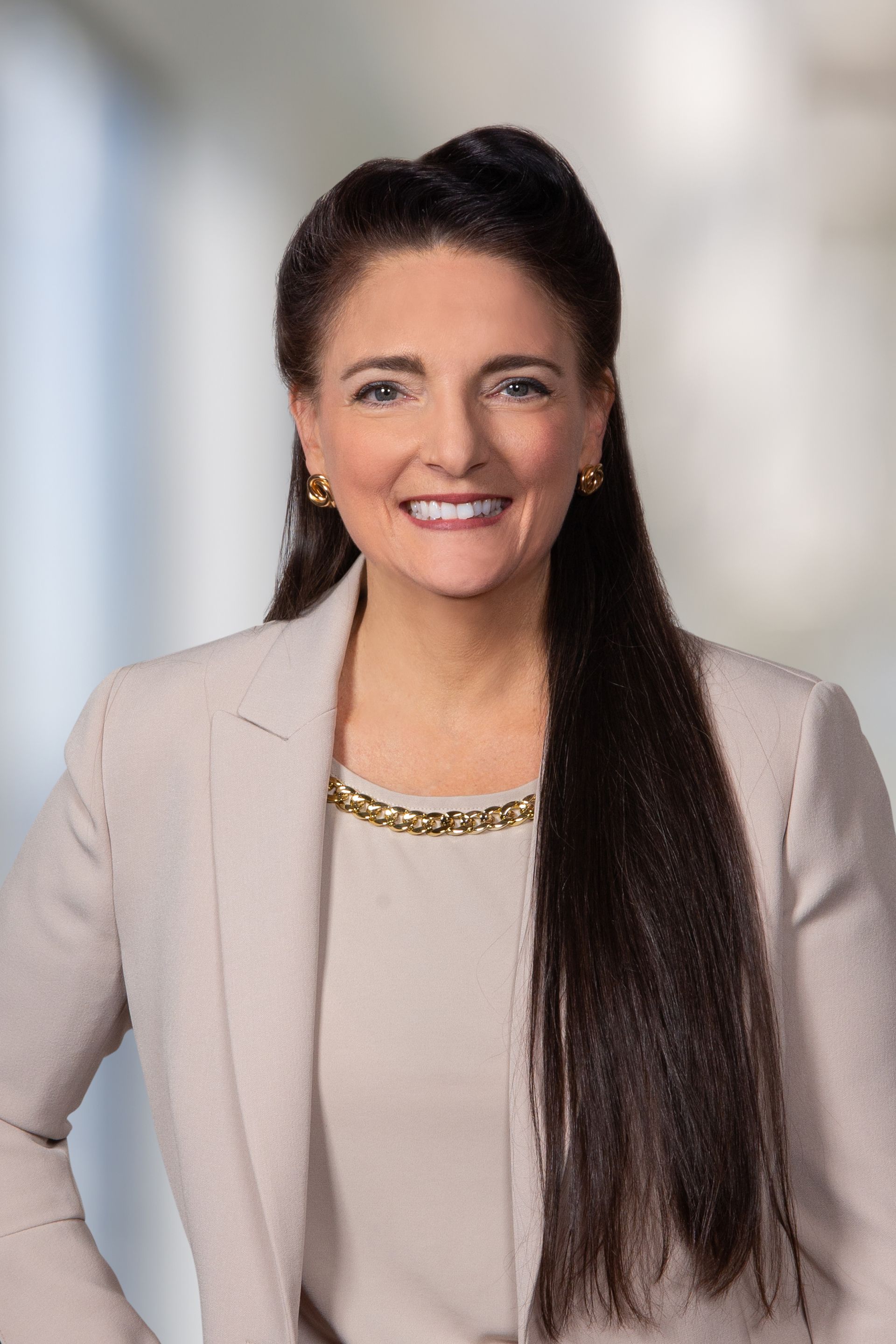 Woman with long dark hair, wearing a light beige blazer and smiling at the camera. Blurred background.