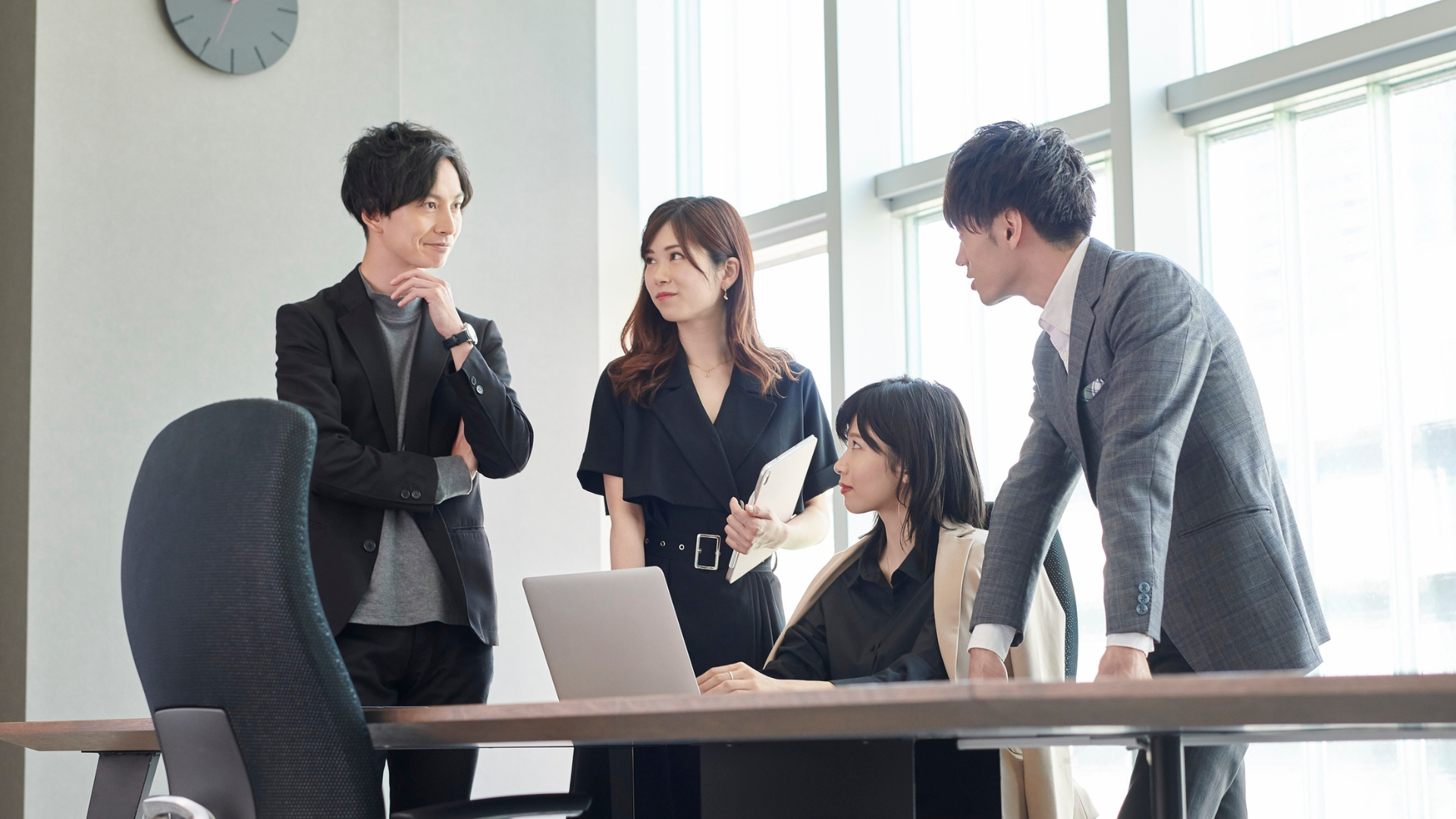 Four colleagues at a desk, reviewing a laptop. One person leans over the desk, others stand. Bright office setting.