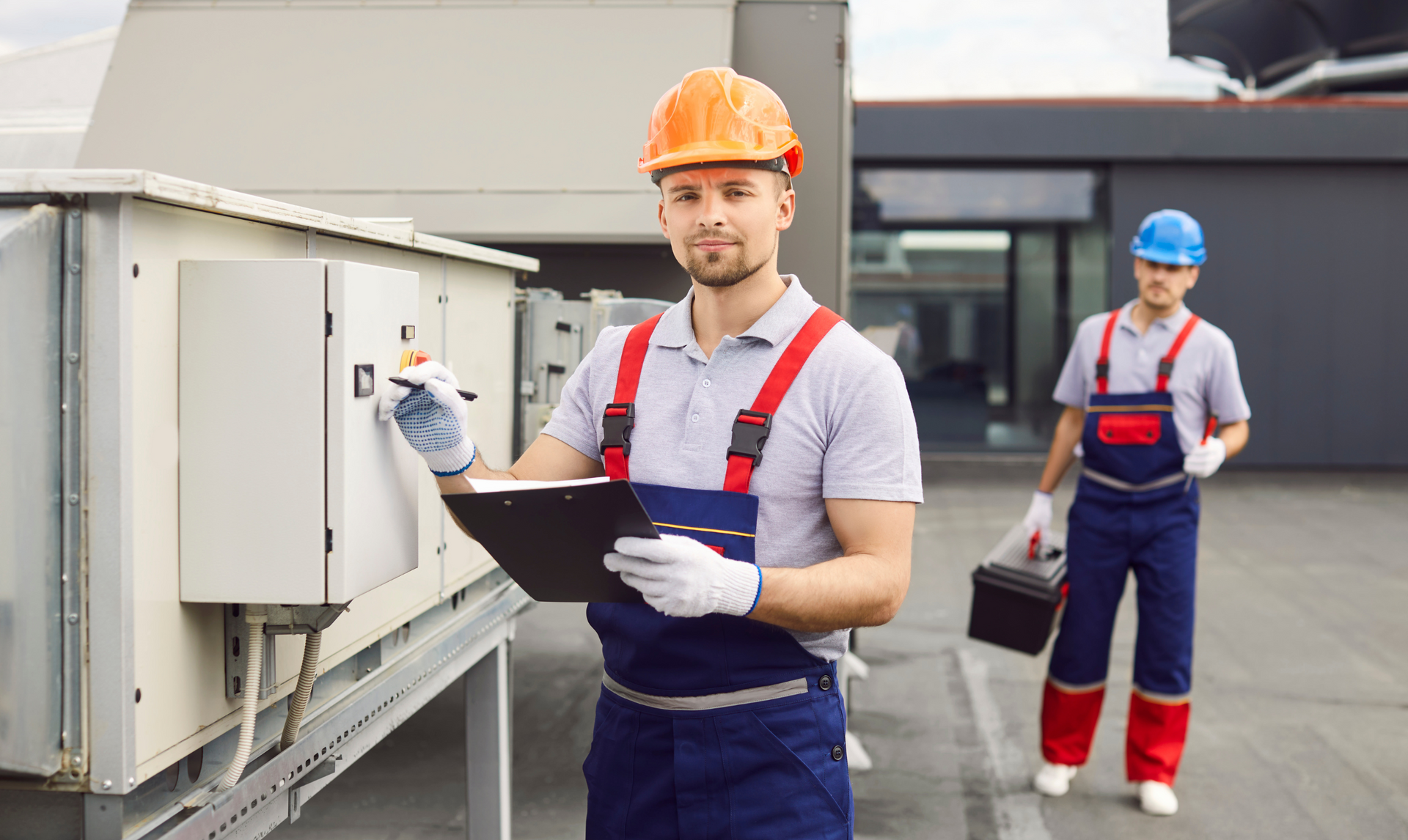 Two HVAC technicians on a rooftop, one taking notes, the other carrying a toolbox.