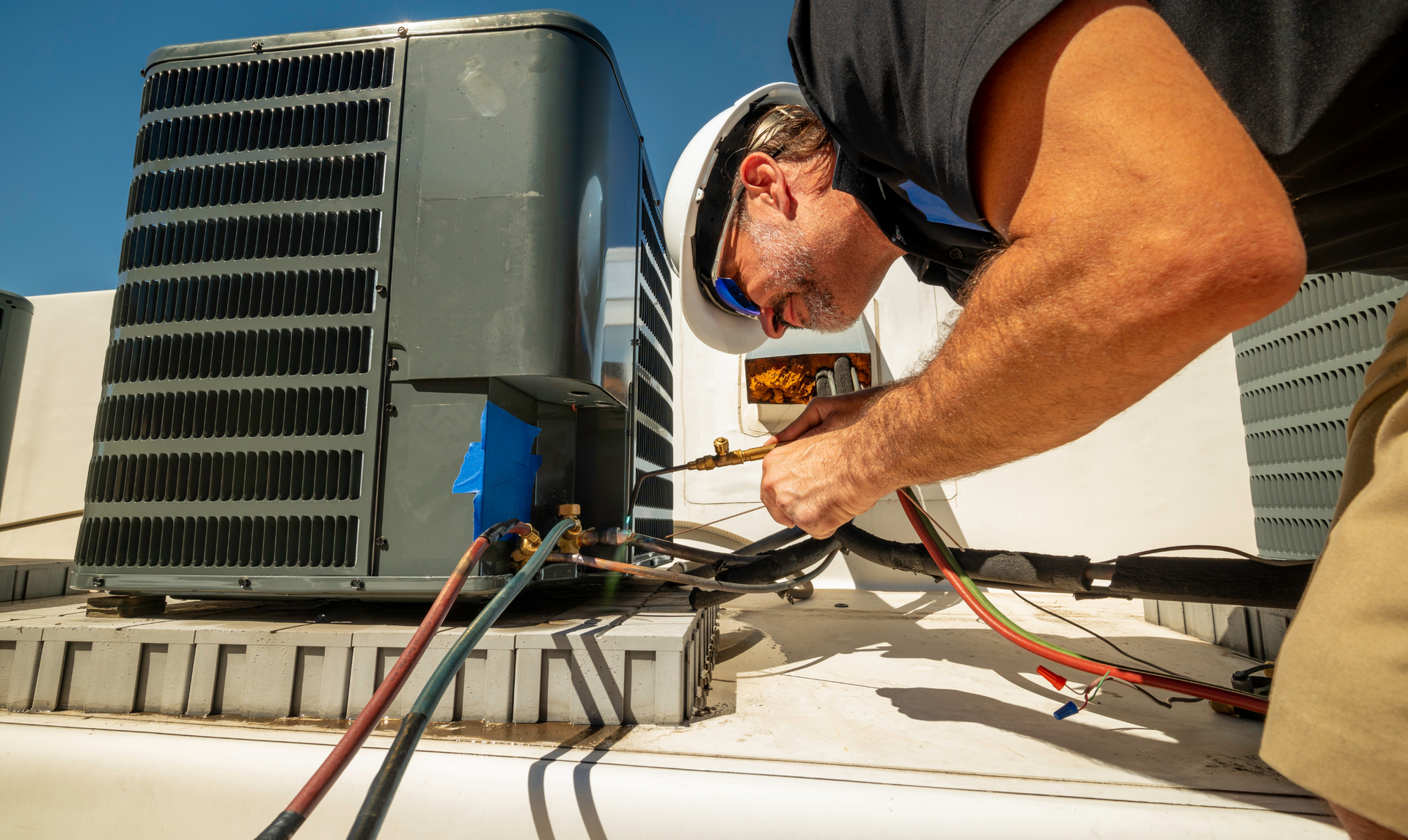 HVAC technician on a rooftop, repairing air conditioning unit with gauges and hoses. Sunny, blue sky.