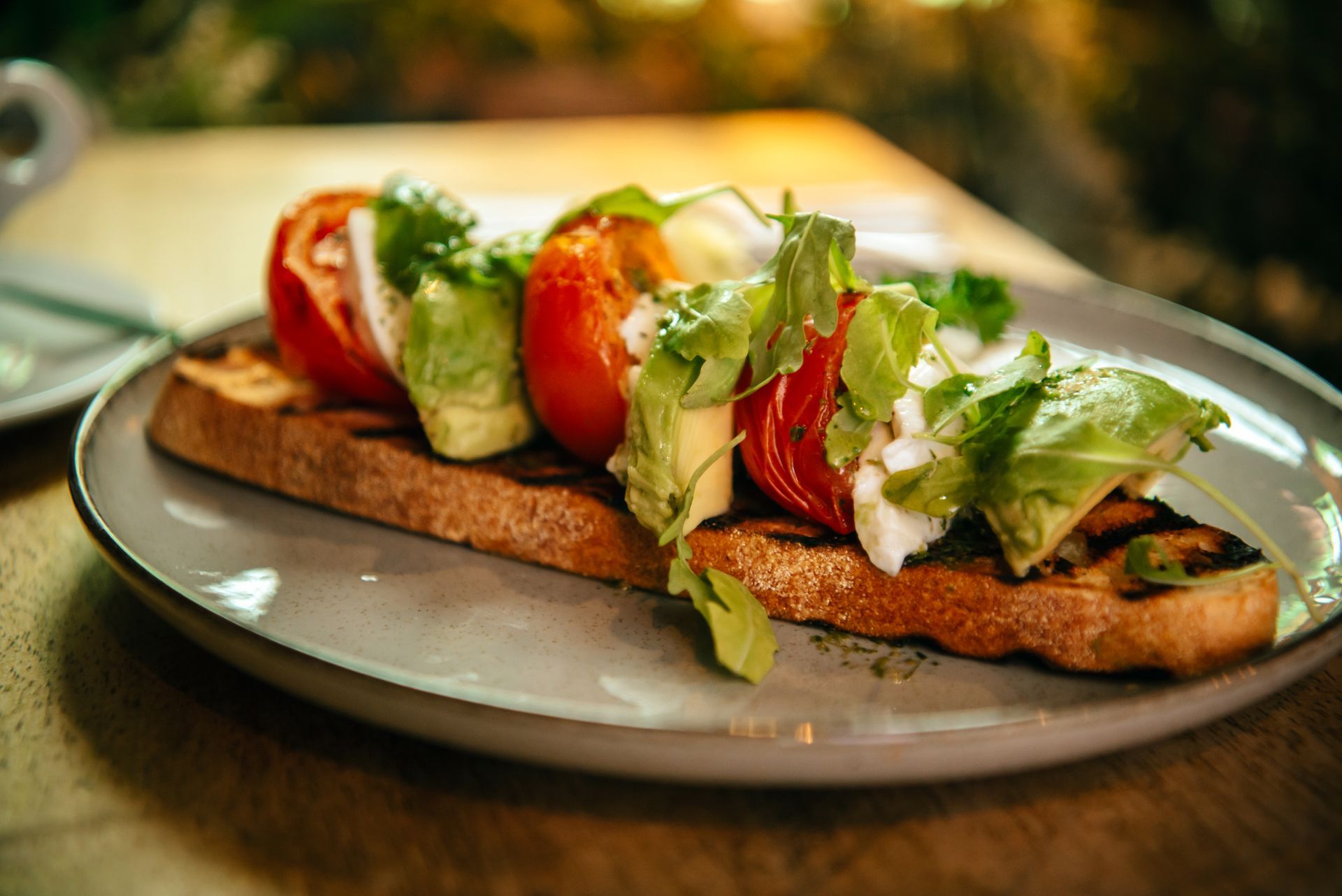 Toast topped with tomato, avocado, arugula, and mozzarella on a plate.