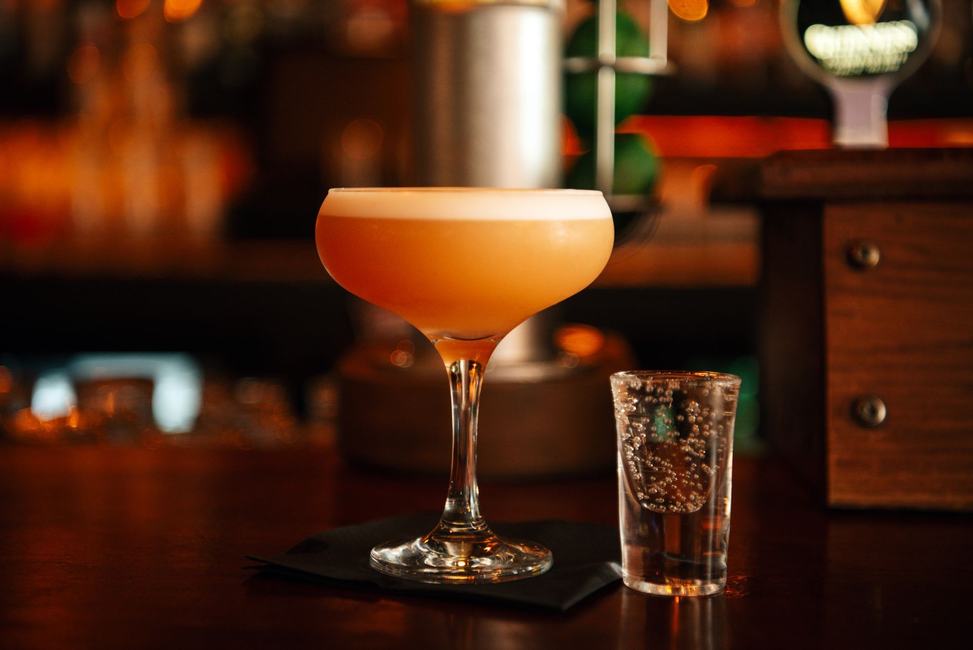 Cocktail in stemmed glass, light orange drink with foamy top, shot glass alongside, bar setting.