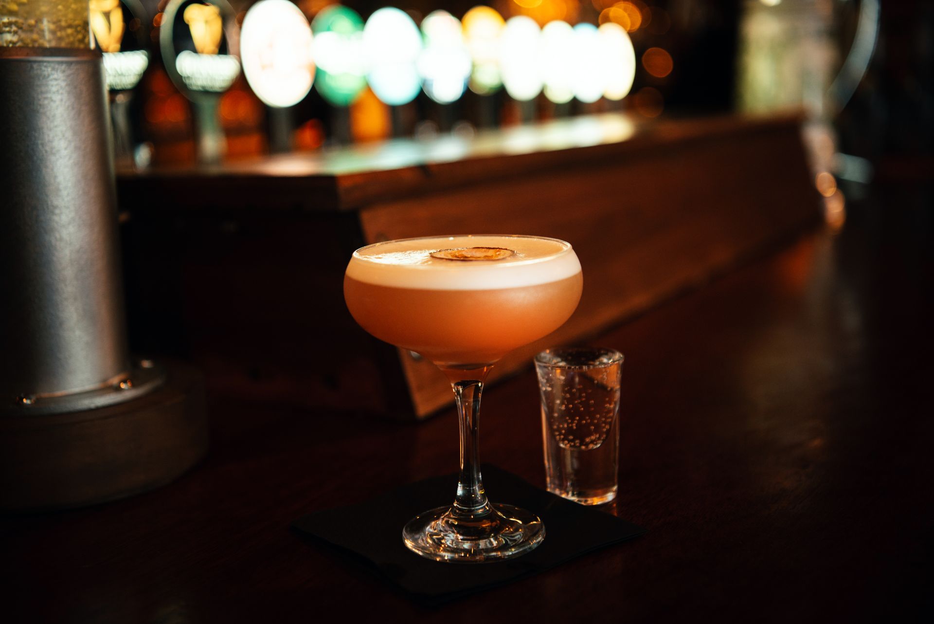 Cocktail with foam in a bar, a small shot glass beside it. The bar is dimly lit.
