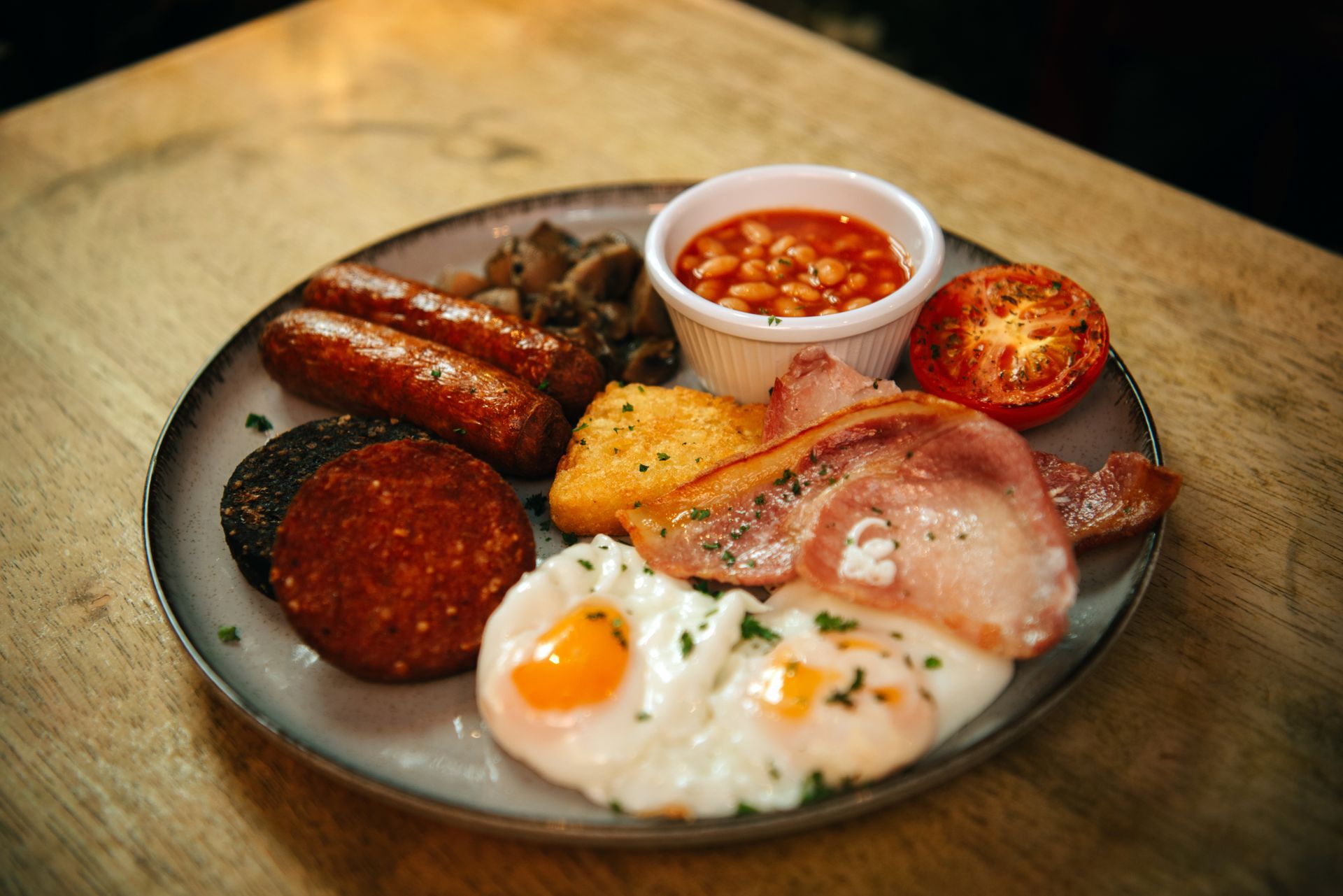 Full English breakfast on a plate: sausages, bacon, eggs, beans, mushrooms, tomato, and black pudding.