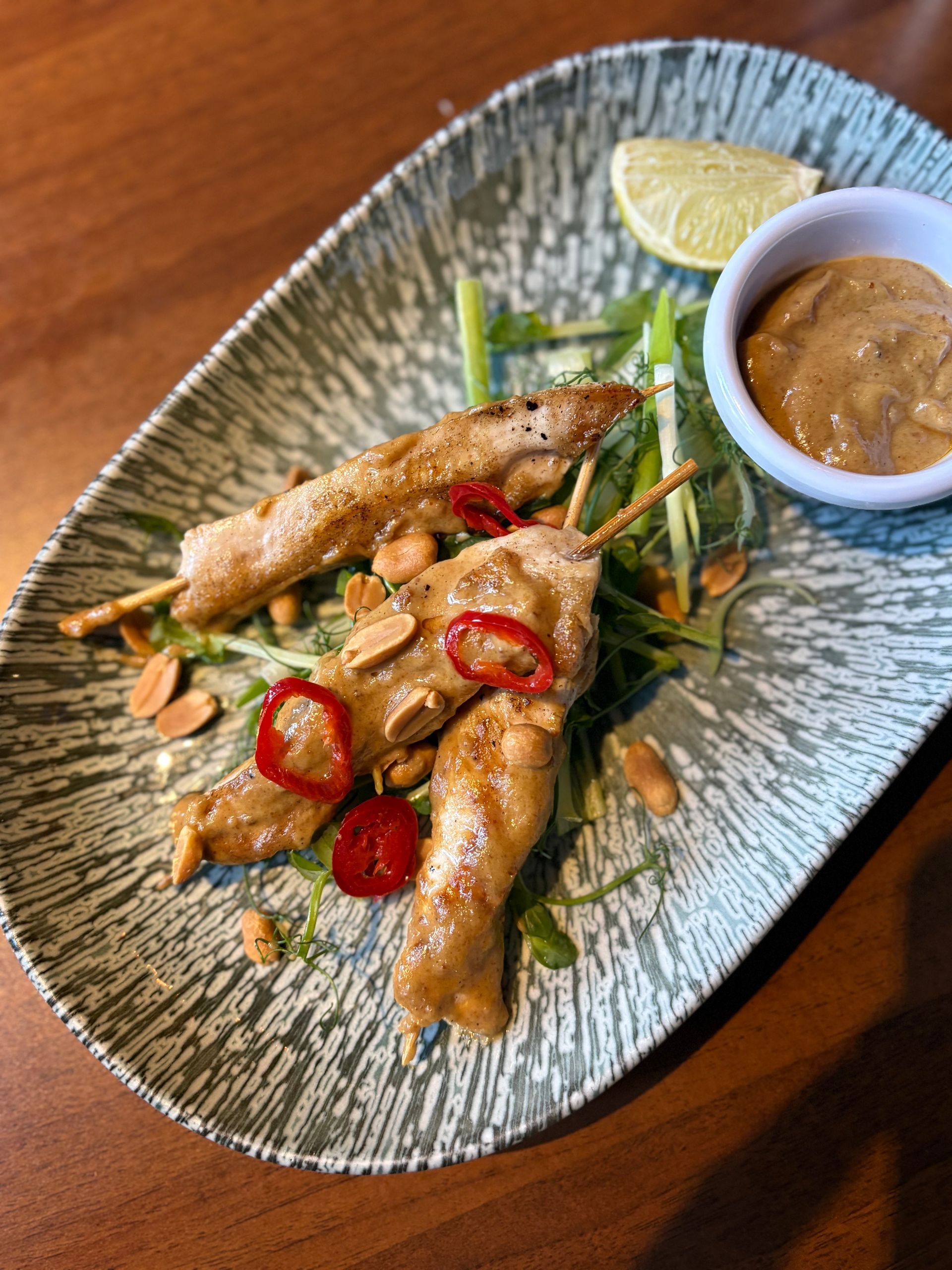 Chicken satay skewers with peanut sauce, chili, and lime on a patterned plate.