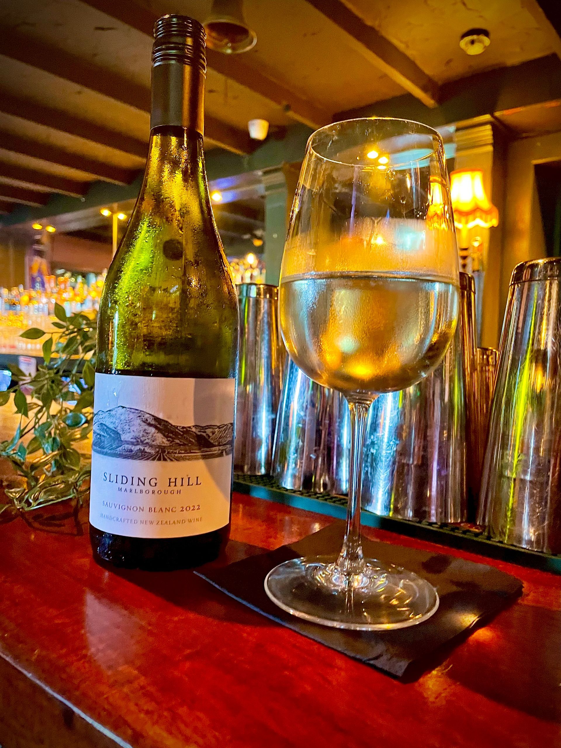 Bottle and glass of white wine on a bar, with cocktail shakers in the background.