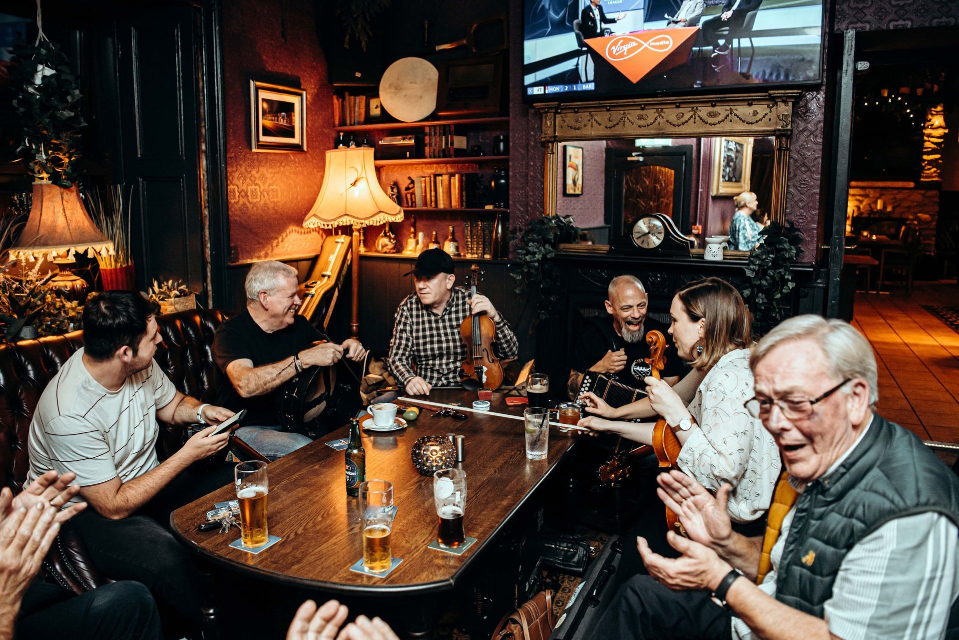 People gathered around a table in a pub, some playing instruments, others clapping and drinking beer.
