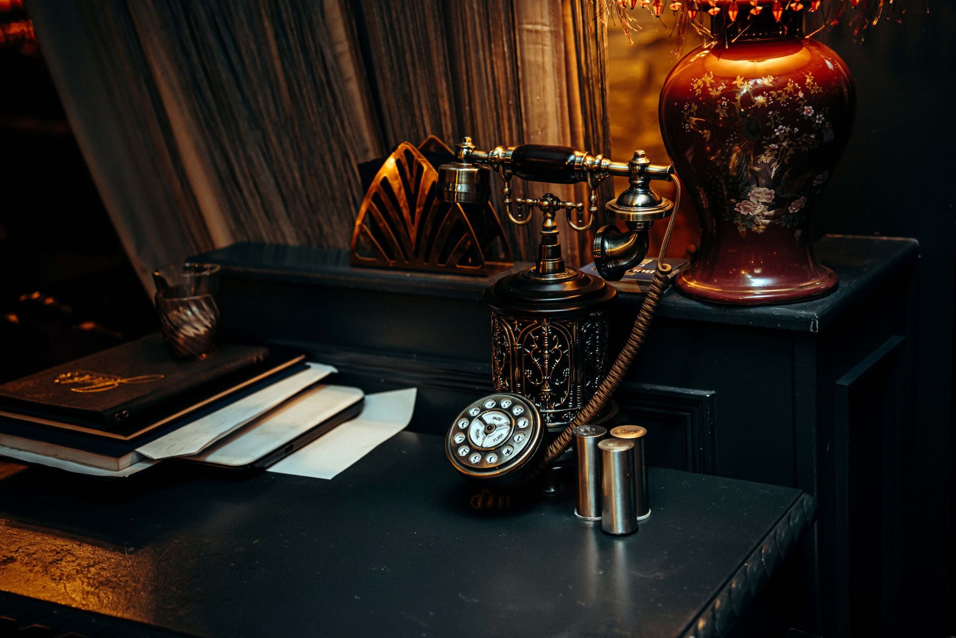 Antique desk with a rotary phone, pocket watch, books, and vase in a dimly lit room.