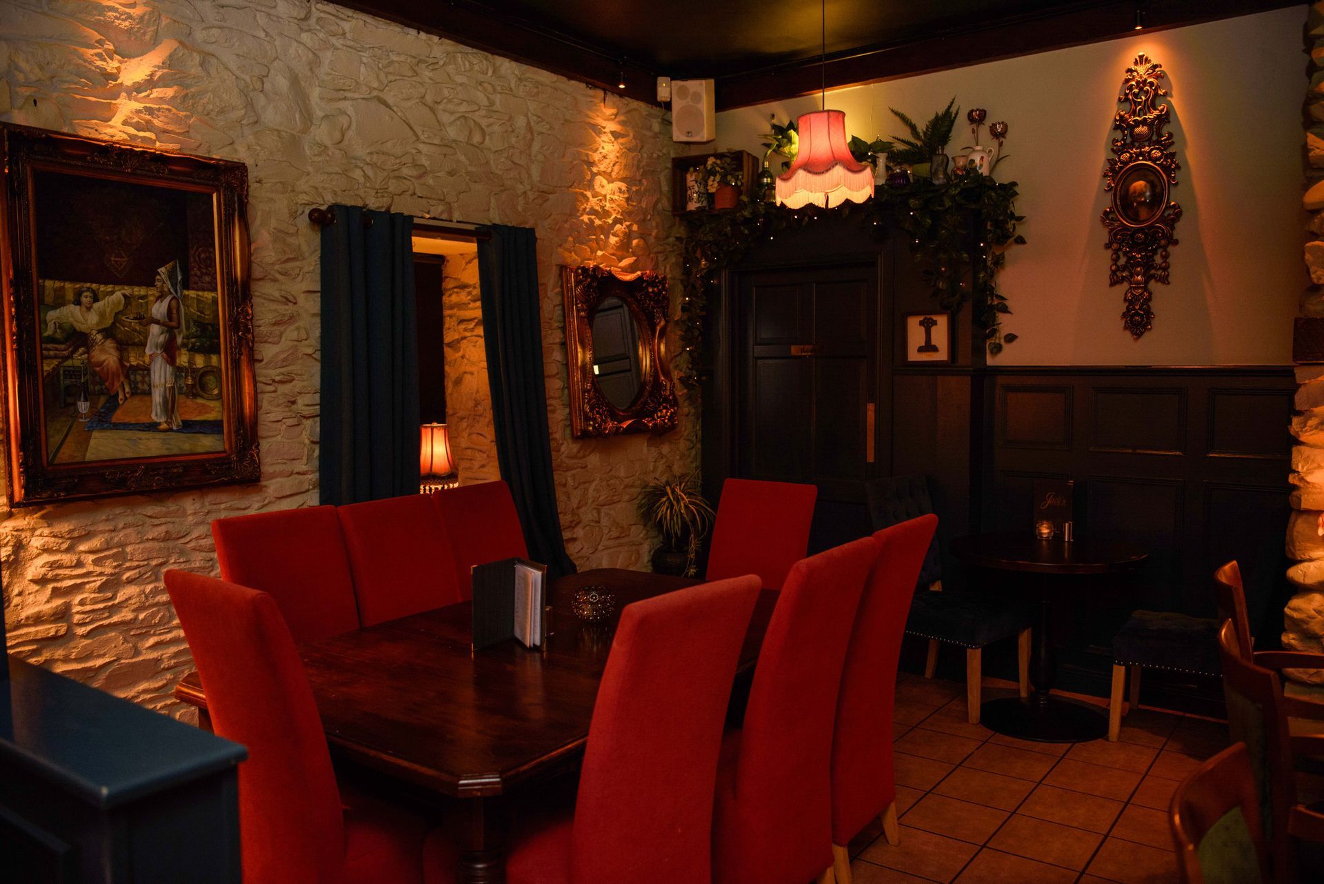 Cozy pub interior with red chairs, dark wood table, stone wall, framed art, and dim lighting.