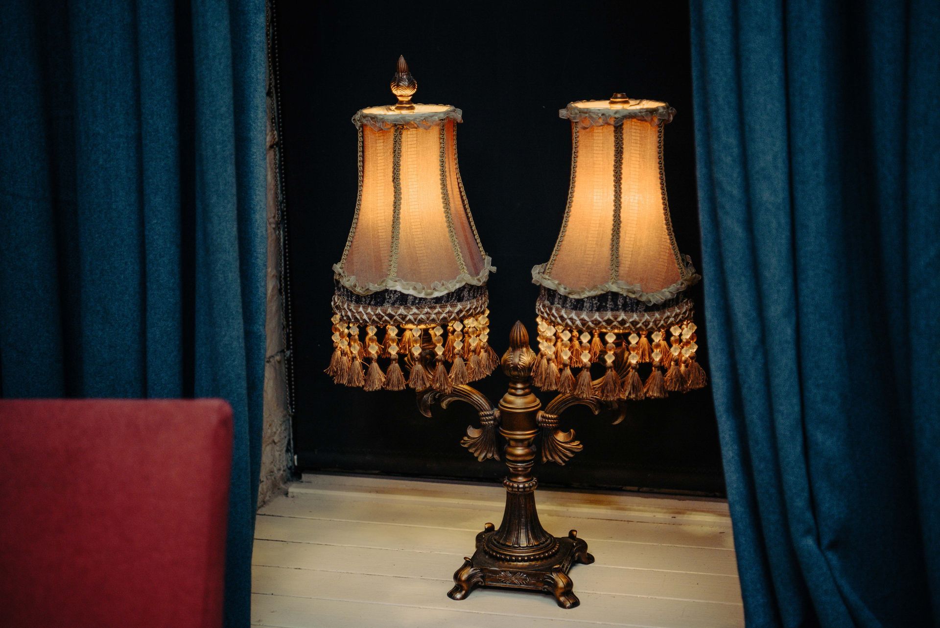 Ornate bronze lamp with two fringed shades, set in a window alcove, dark blue curtains.