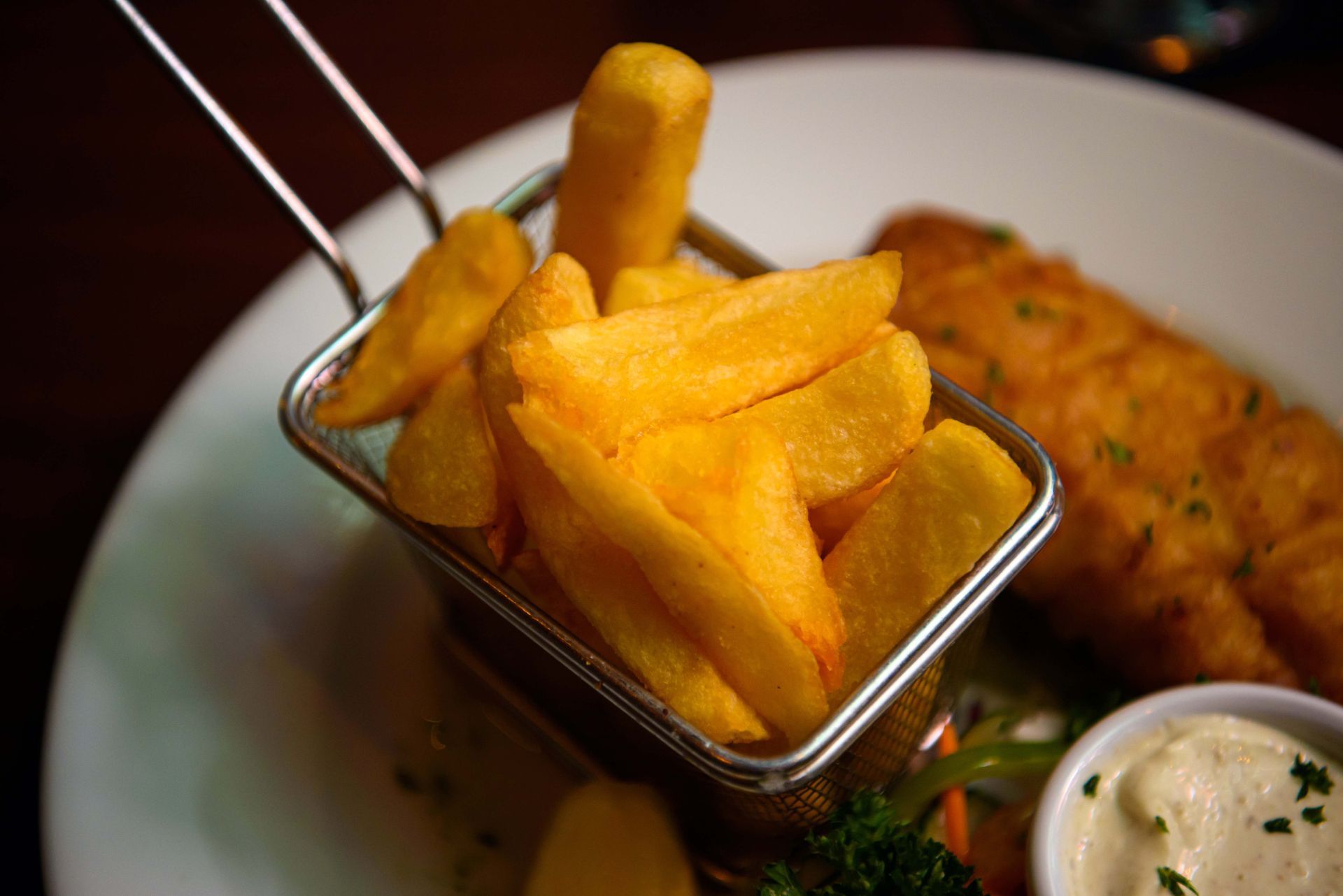 Golden fries in a wire basket, with fried fish, vegetables, and sauce on a white plate.