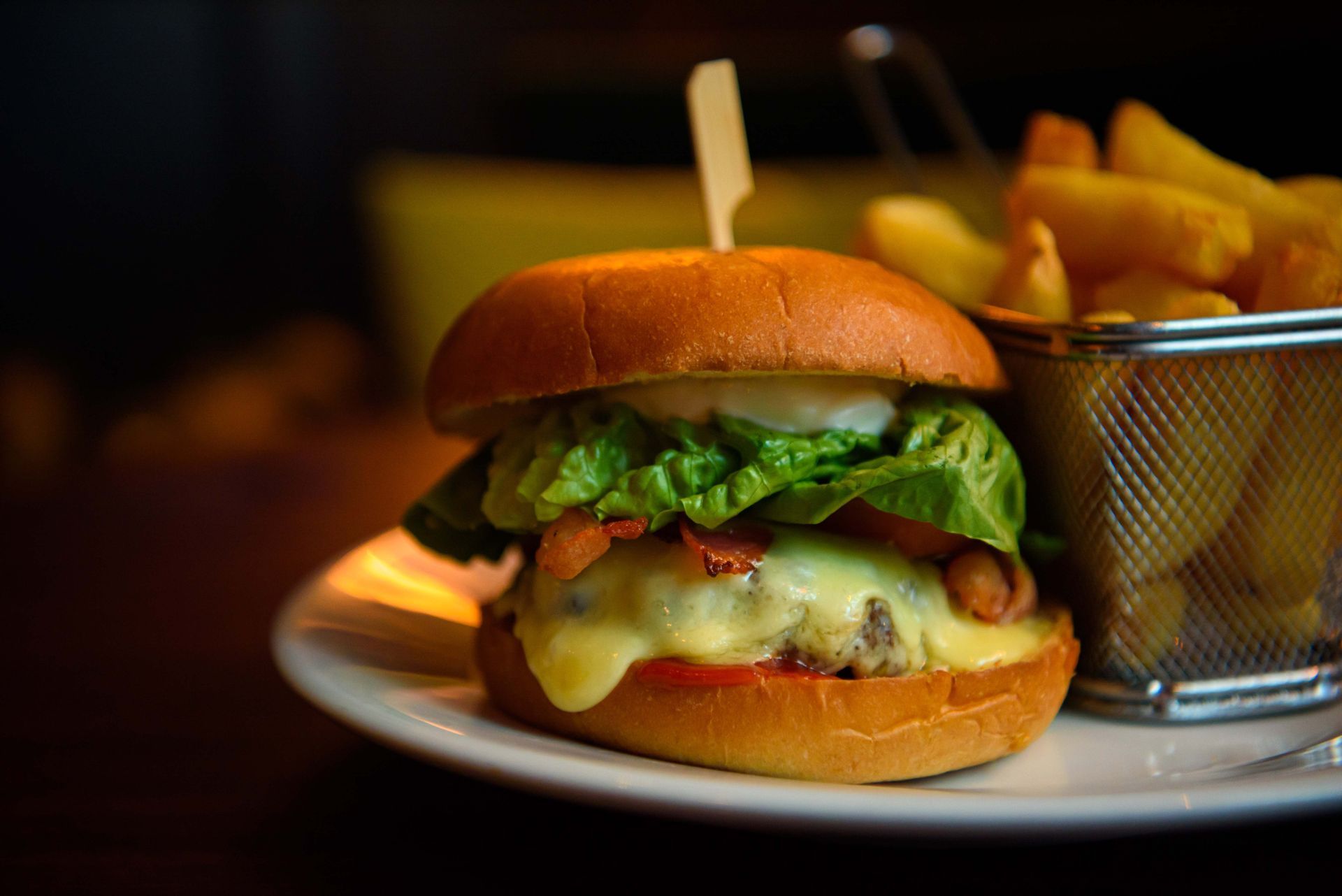 Hamburger with lettuce, cheese, and bacon; fries in a wire basket on a white plate.