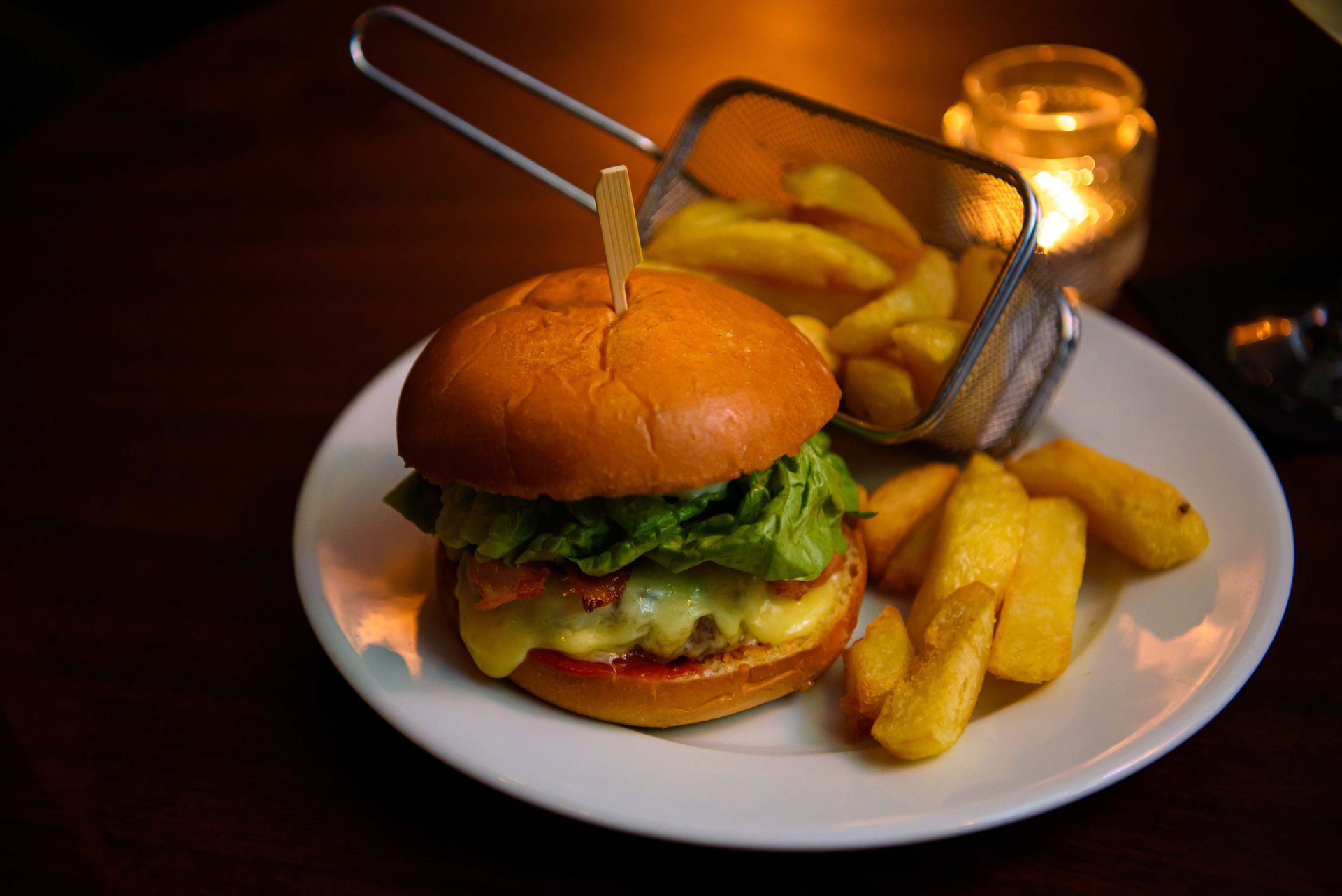 Hamburger on a plate with fries and a metal basket, with a candle in the background.