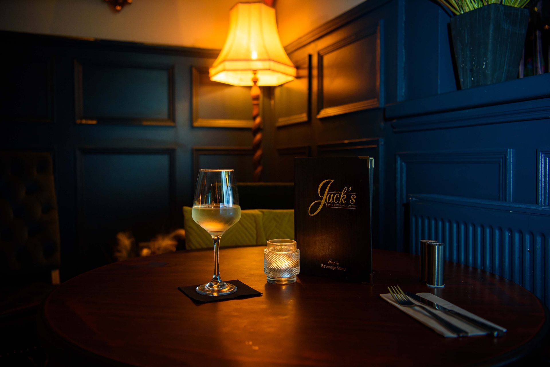 Cozy restaurant table set for one, with a wine glass, menu, and candle, under a warm lamp.