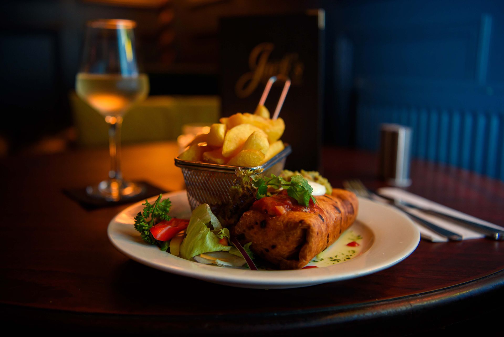Plate of food with fries, a glass of wine, and menu on a dark table.