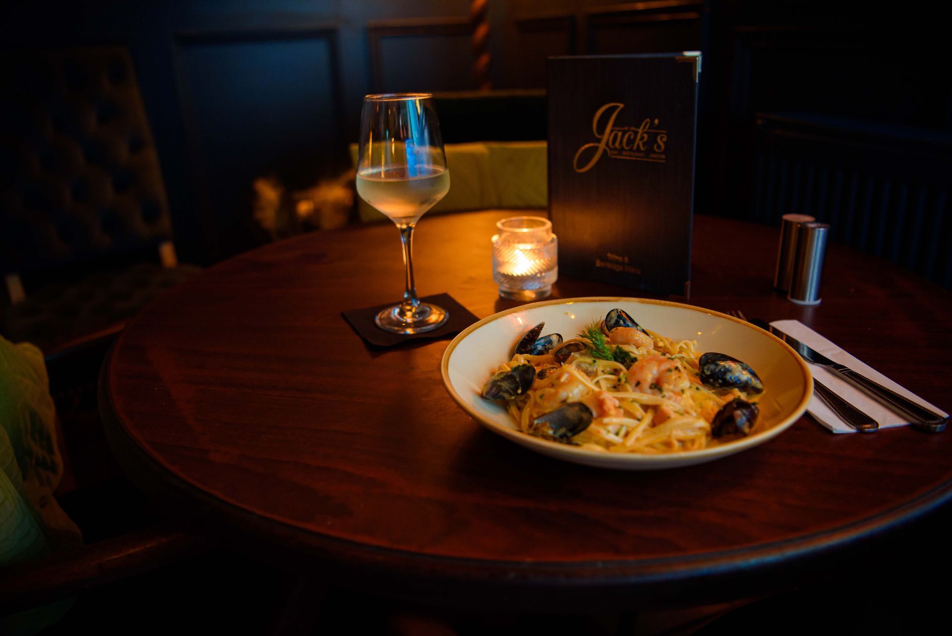 Pasta with mussels, wine glass, candle, and menu on a dark wooden table.