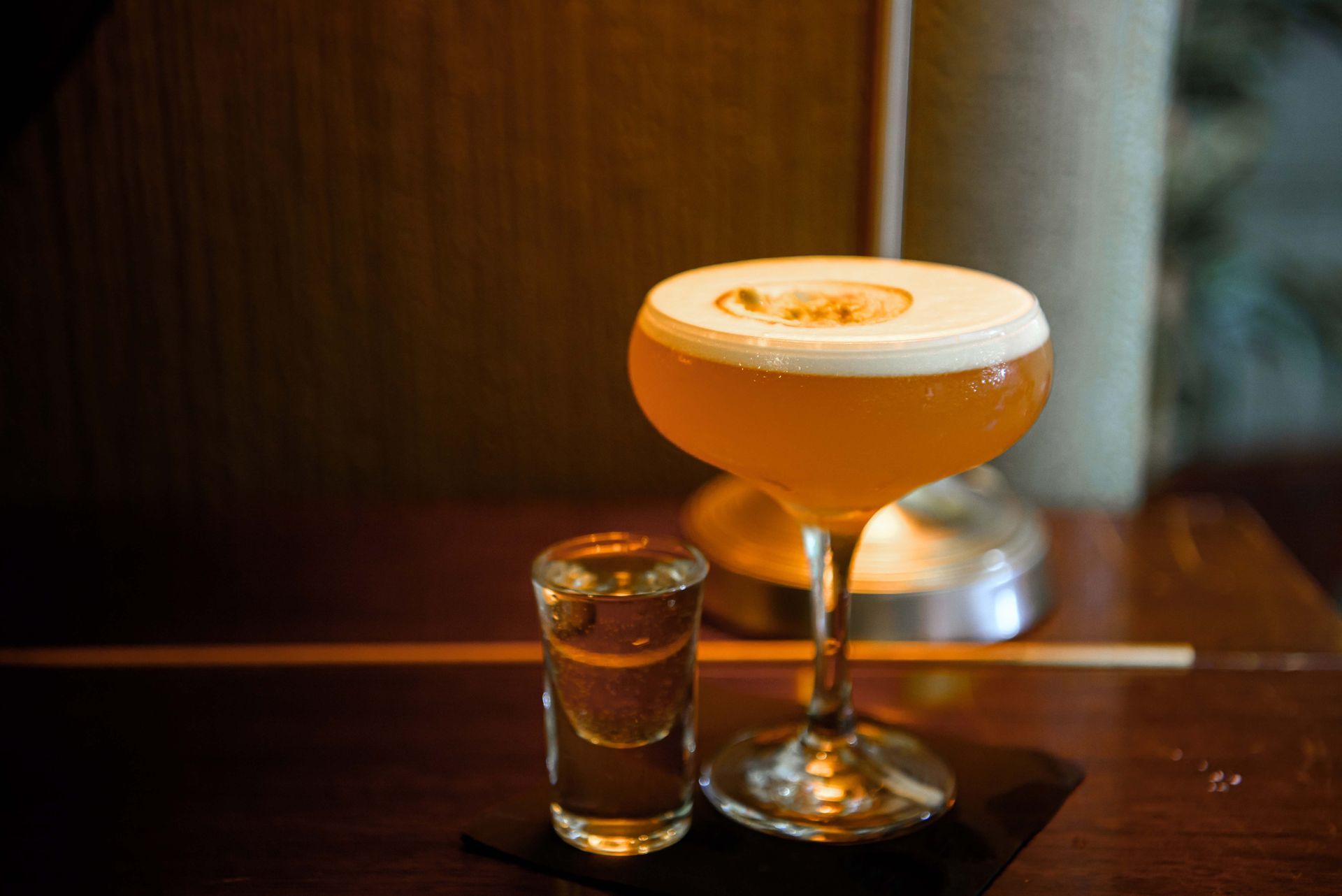Coupe glass of golden cocktail with foam, gold leaf, shot glass on a wooden bar.