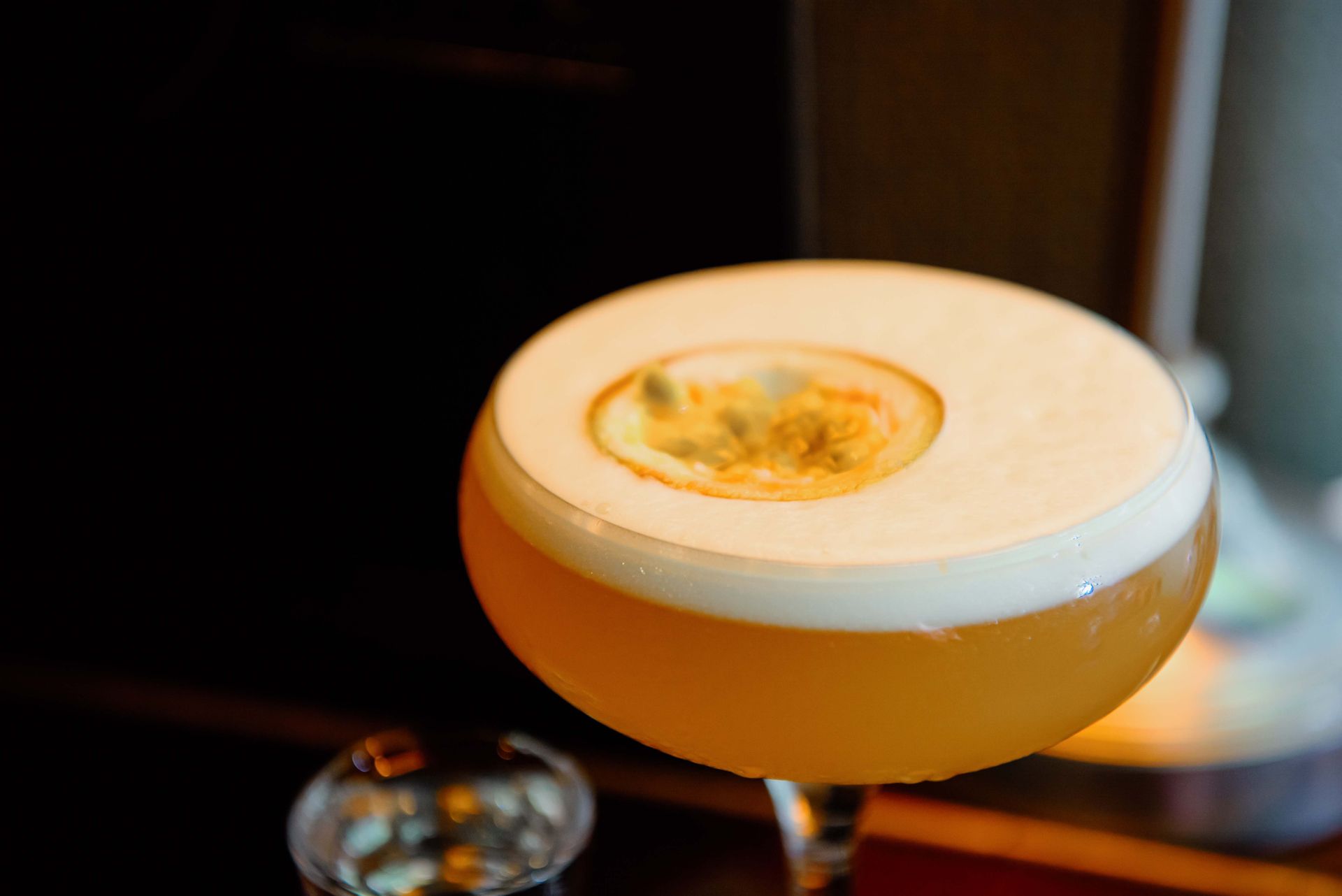 Cocktail in a stemmed glass with a frothy white top and passion fruit garnish.