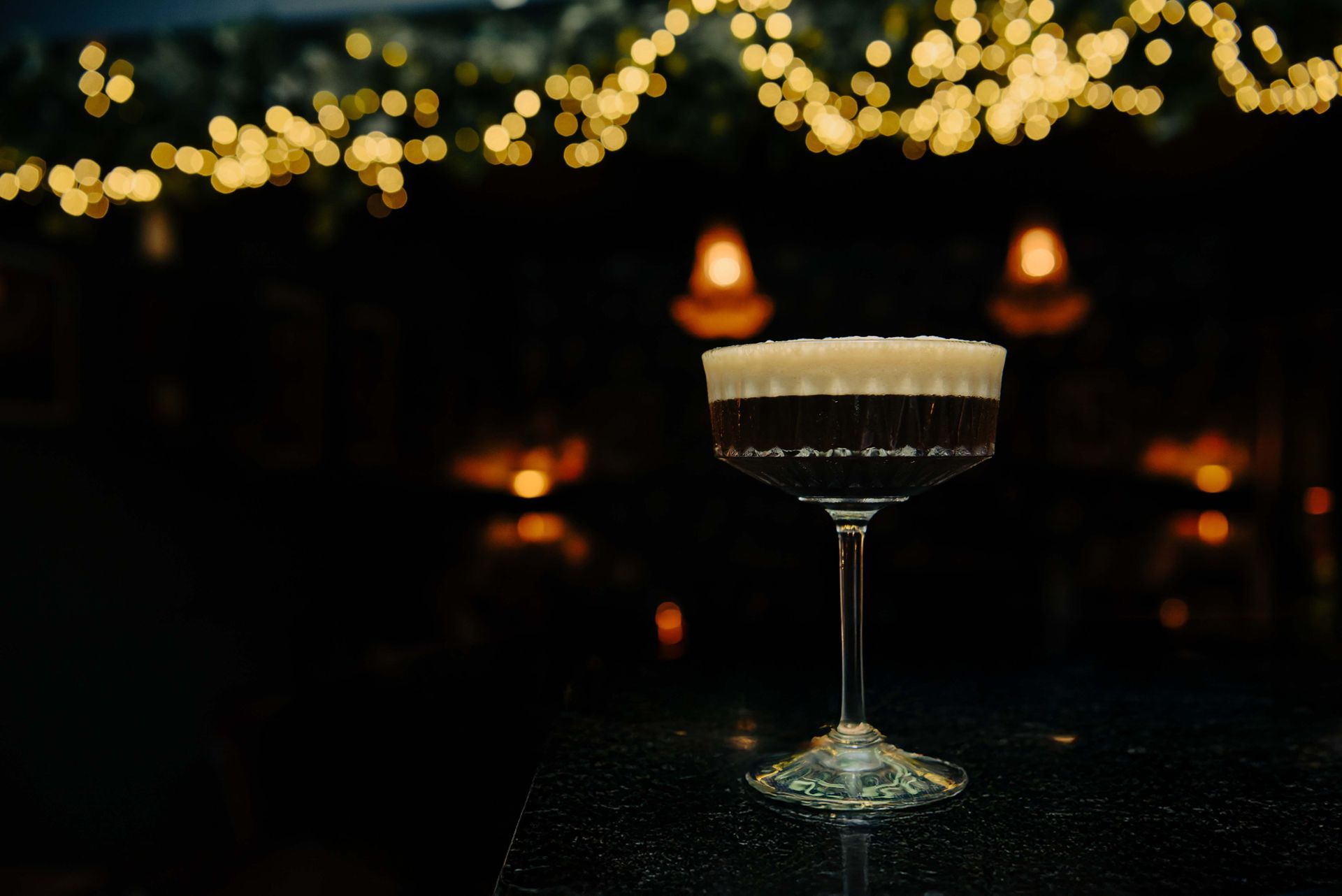 Cocktail in a stemmed glass with a dark liquid base and foamy white top; set against blurred bokeh lights.