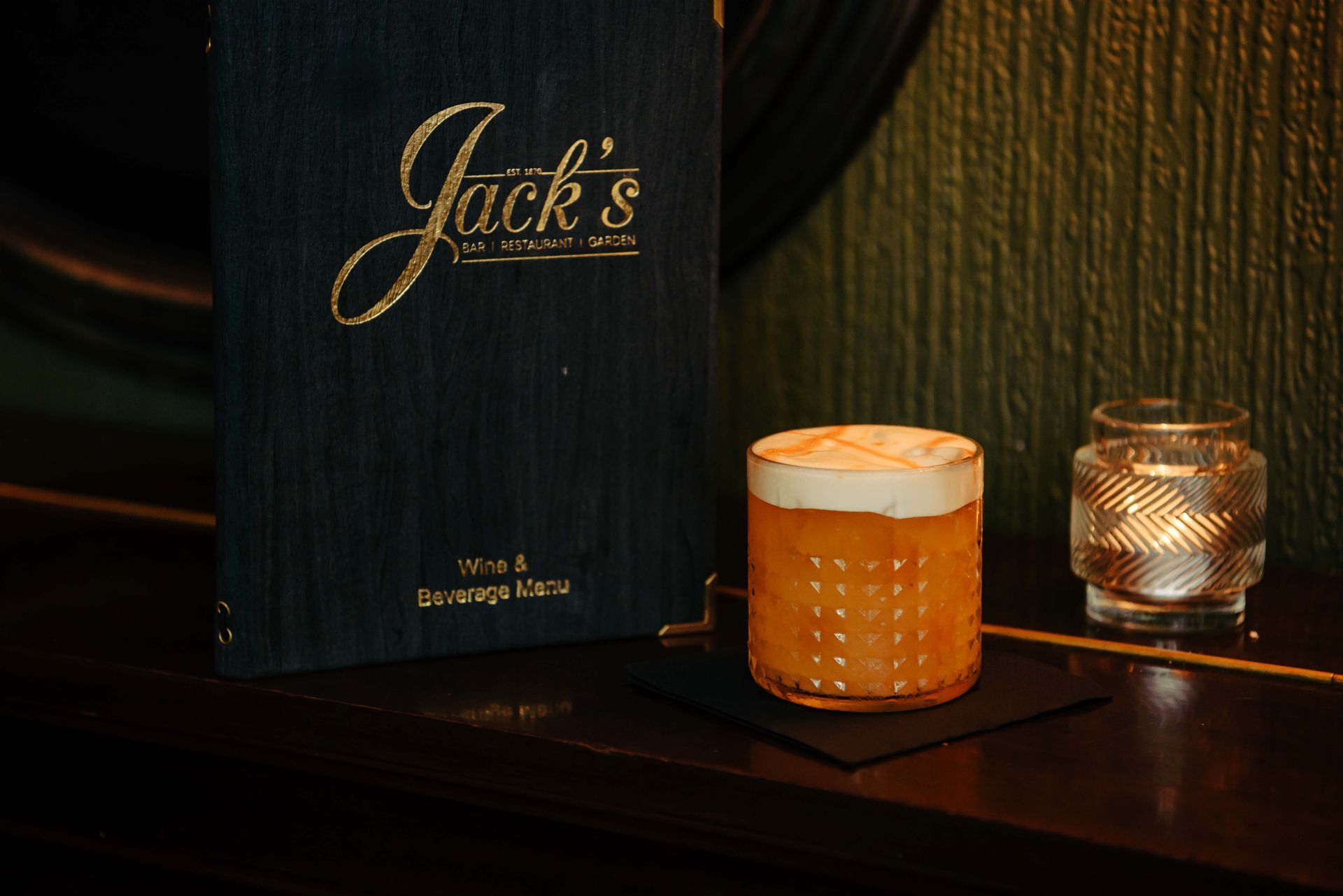 Cocktail and menu at Jack's Bar. Orange drink in a glass with foam, lit candle, and black menu.