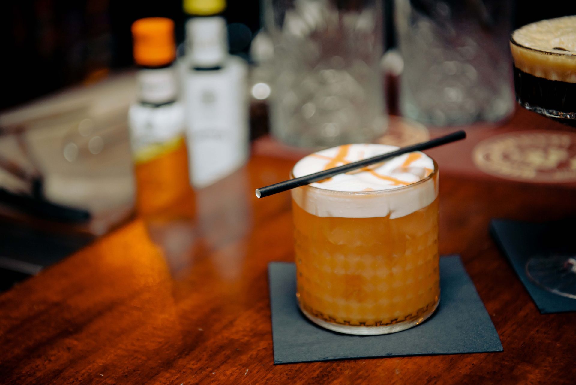 Cocktail with white foam top and caramel drizzle, black straw, on a coaster, on a wooden bar.