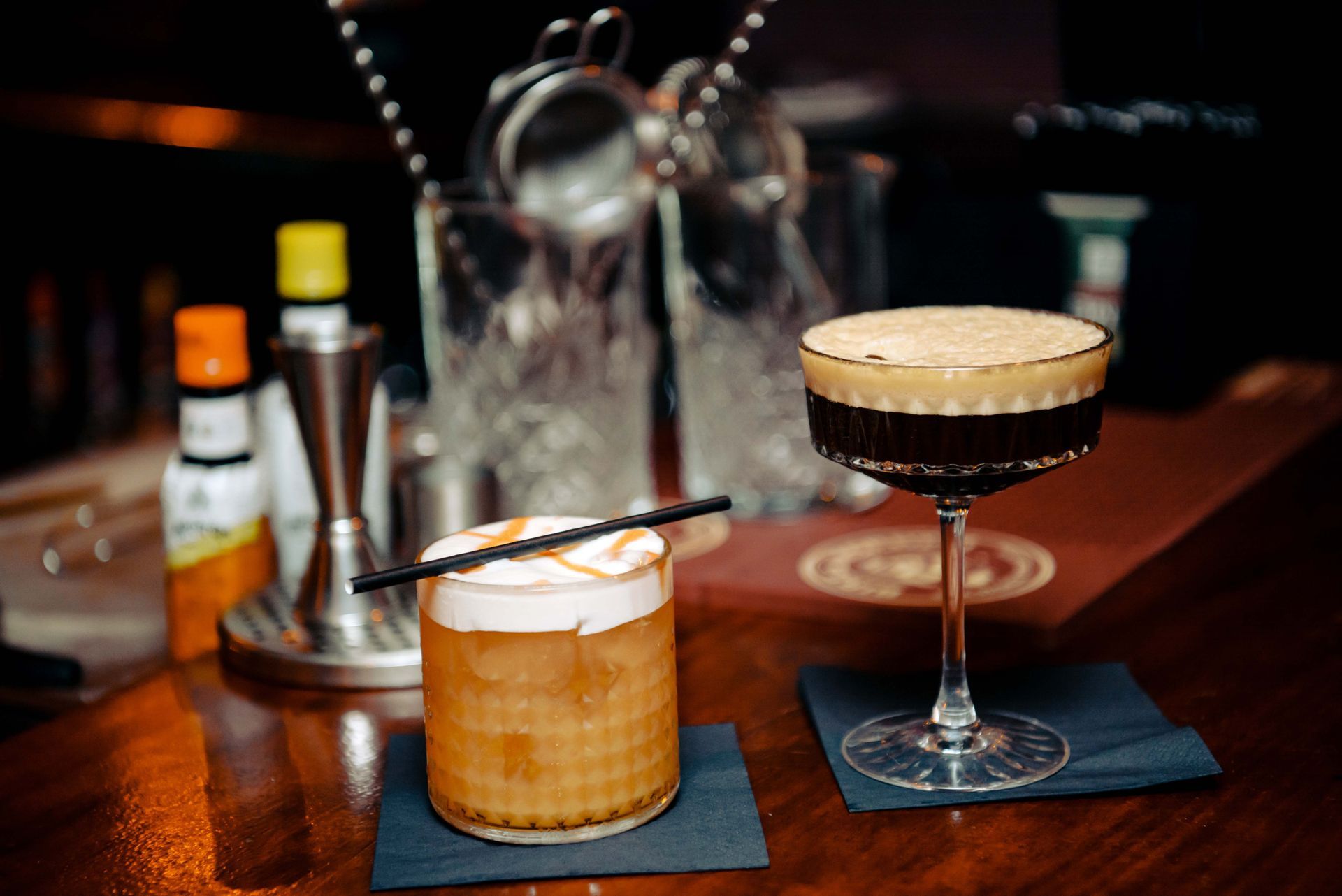 Two cocktails on a bar; one with foam and garnish, the other dark with foam. Bar tools in background.