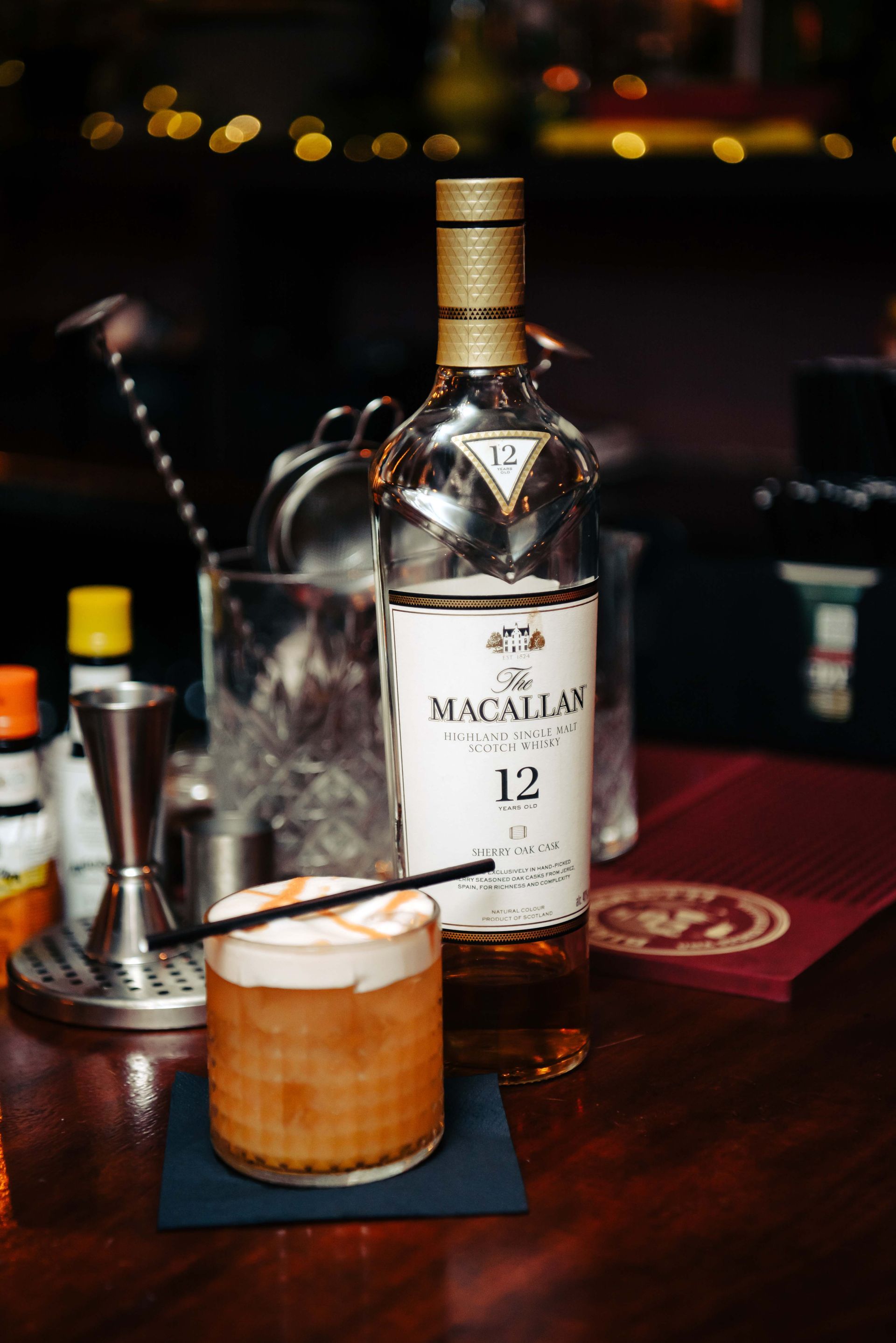 Cocktail with foam topping next to a bottle of The Macallan 12 whiskey on a bar.