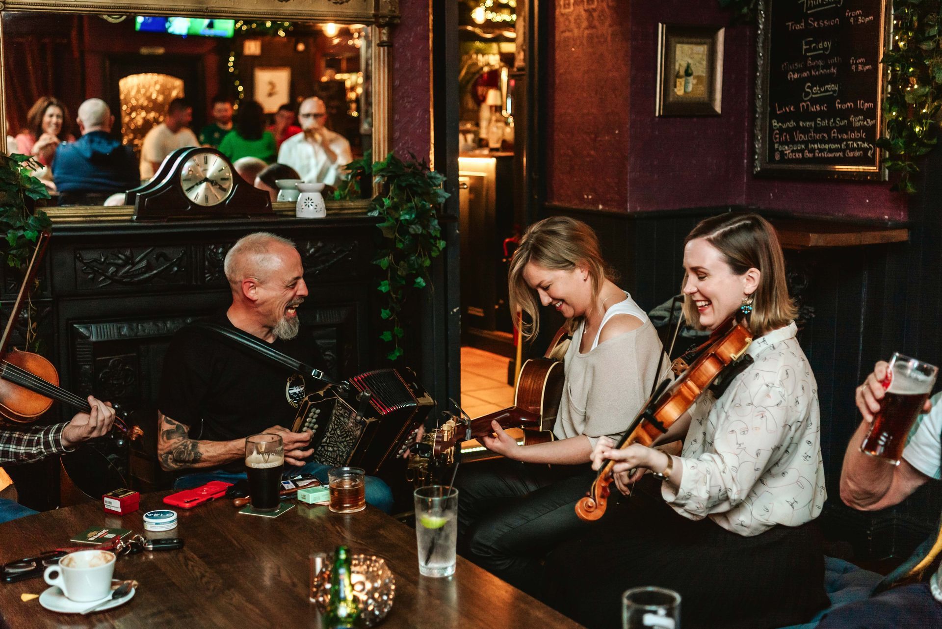 Musicians play string instruments in a pub with an audience. They are smiling.