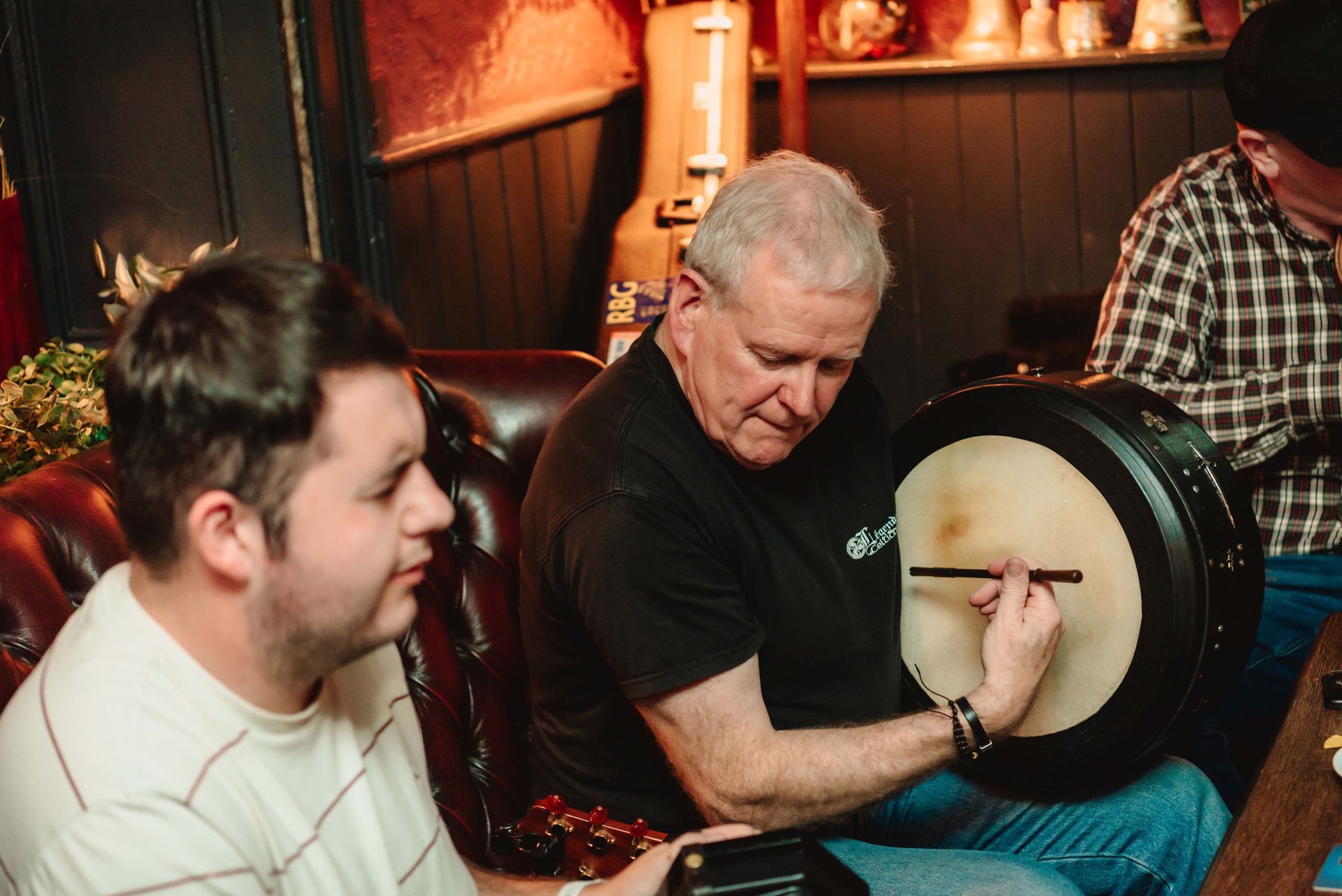 Man plays the bodhrán, accompanied by two others in a dimly lit setting.