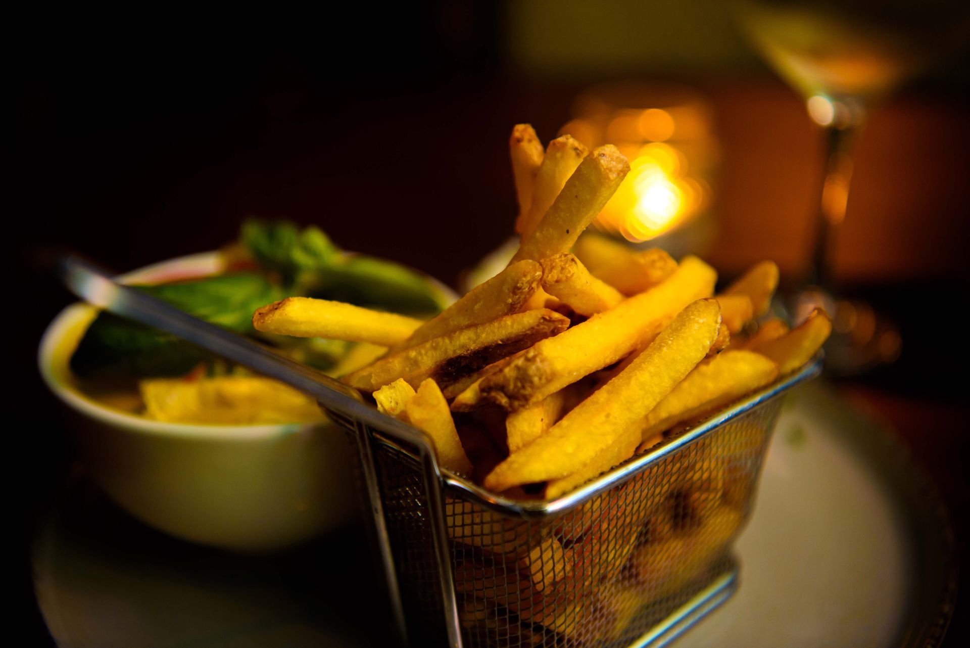 Golden french fries in a metal basket with dipping sauce, candlelit setting.